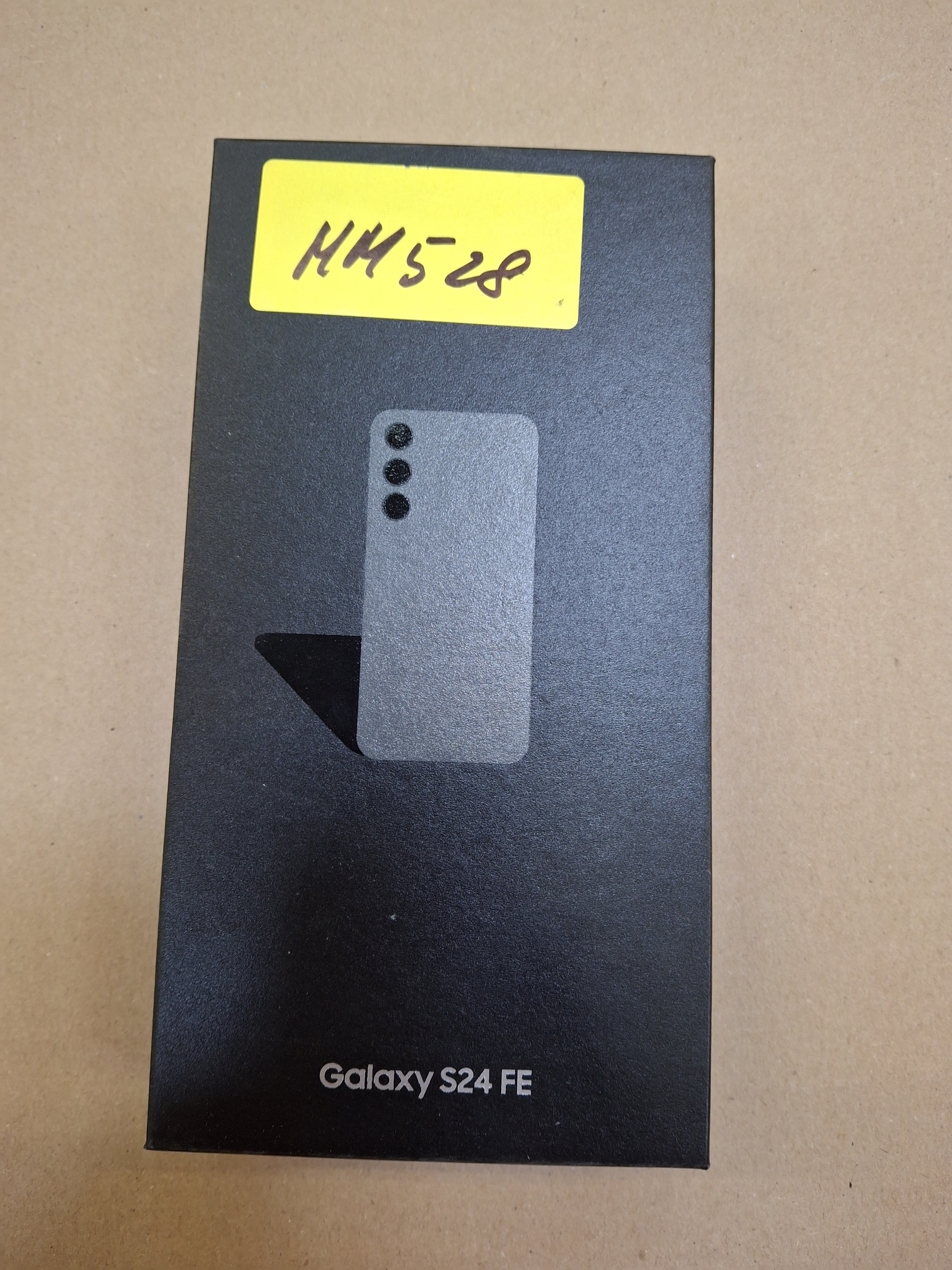 Czarne pudełko Galaxy S24 FE z obrysem telefonu i żółtą naklejką. Tło to brązowa powierzchnia.