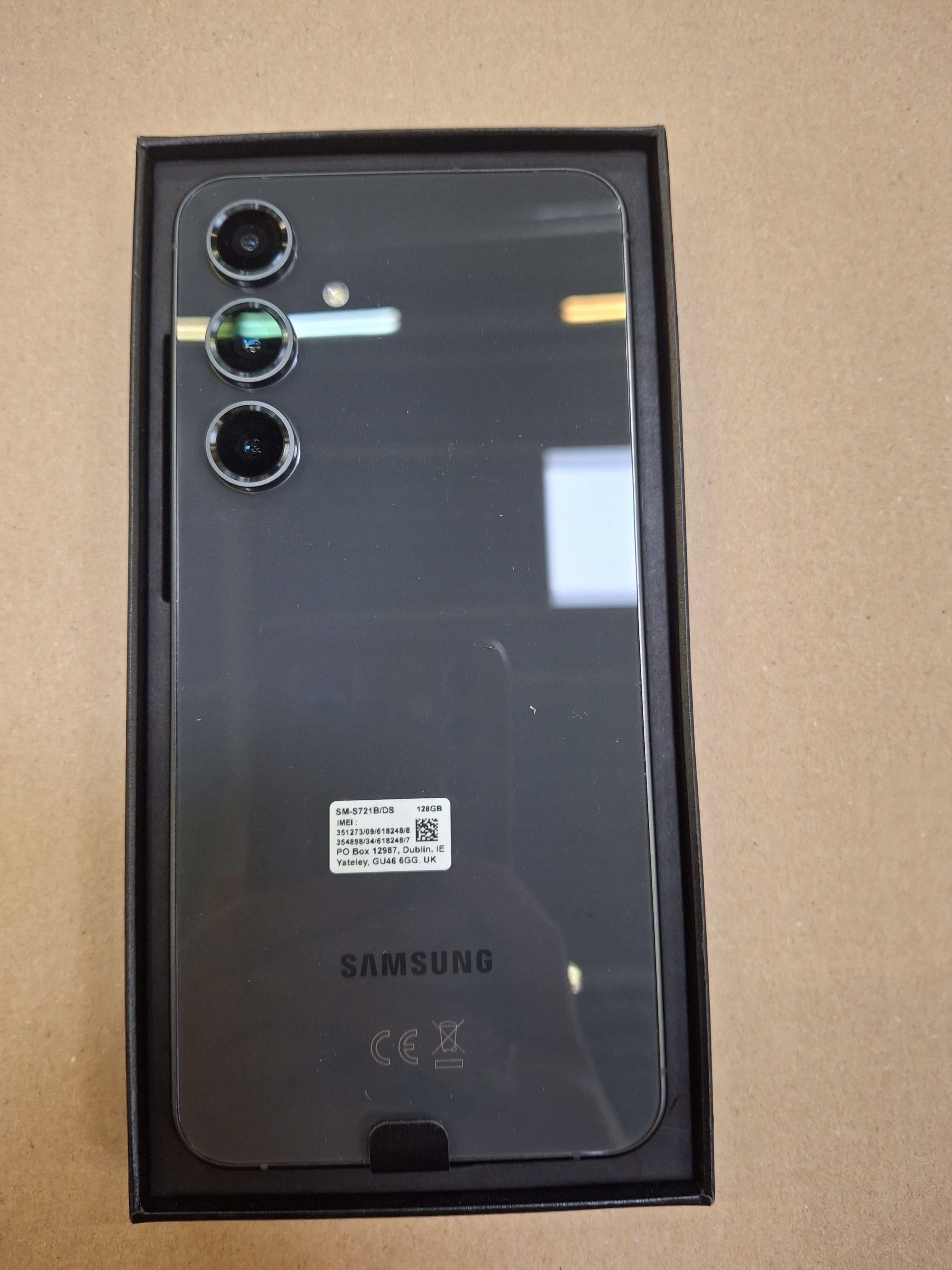 Otwarta czarna skrzynka na telefon z szarym telefonem. Telefon ma trzy aparaty i logo Samsung. Brązowe tło.