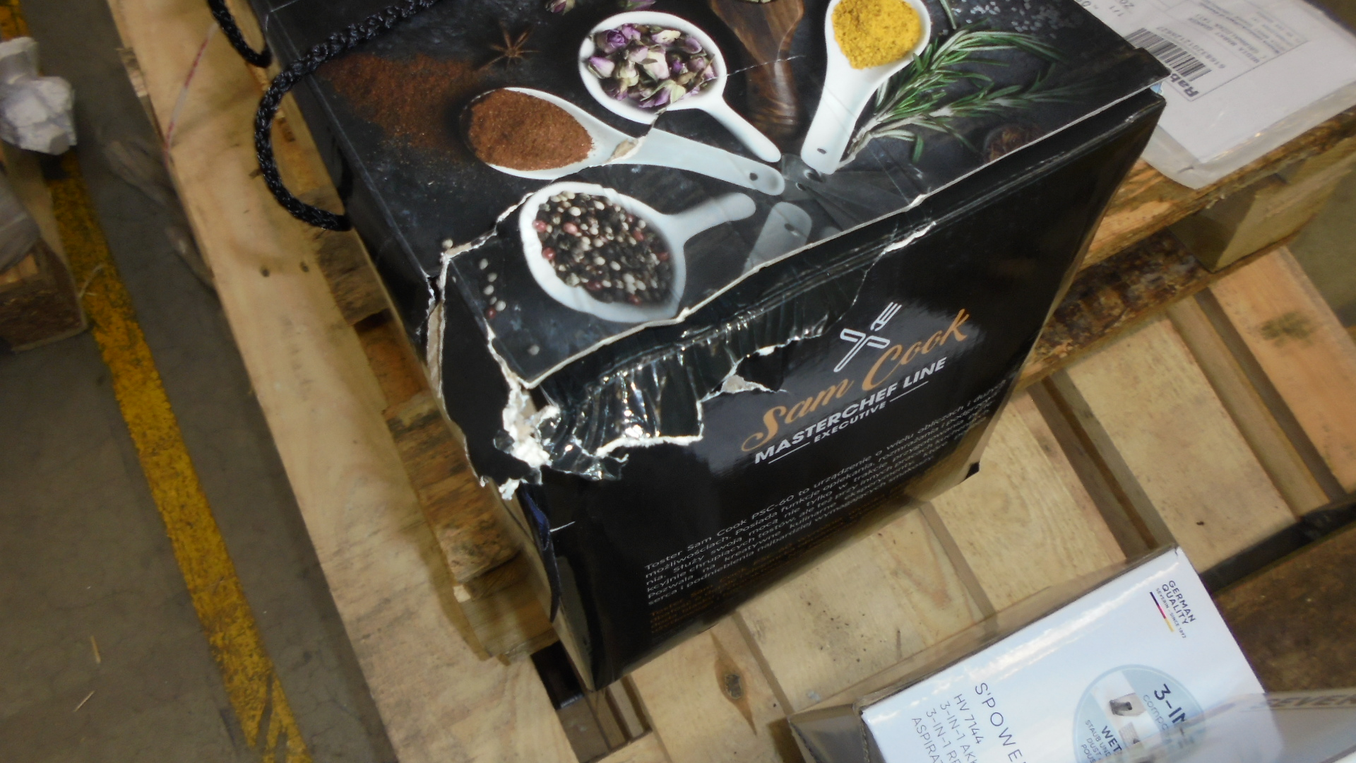 Uszkodzone pudełko Sam Cook z przyprawami na łyżkach. Pudełko jest na palecie, w tle widoczny inny pakunek.