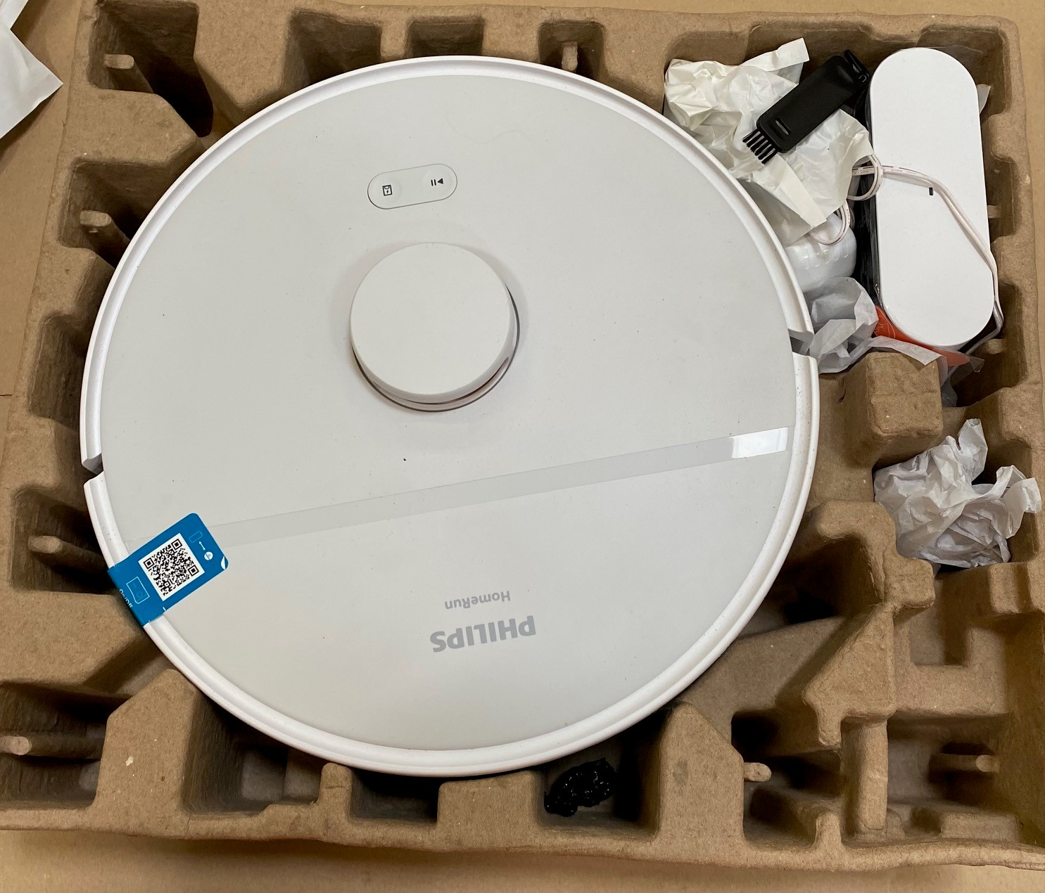 Biały robot odkurzający Philips HomeRun wewnątrz kartonowego opakowania. Akcesoria obok. Widok z góry, zbliżenie.