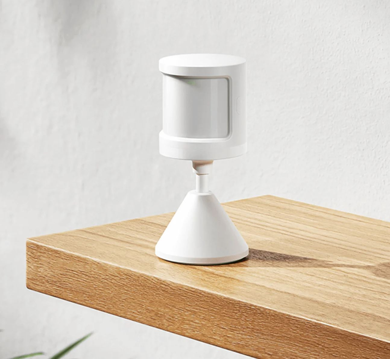 Biały czujnik ruchu na drewnianej powierzchni. Czujnik ma cylindryczną górę i stożkową podstawę. Tło to biała ściana.