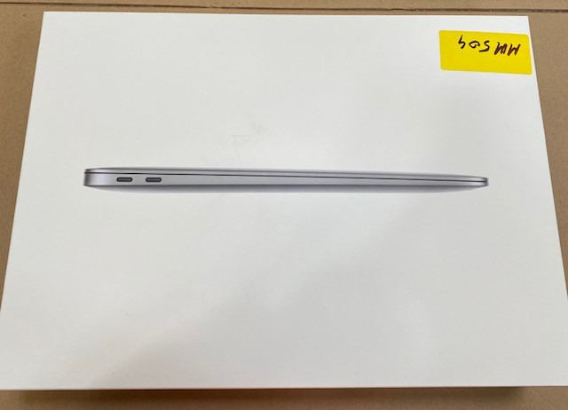 Widok z boku srebrnego MacBooka w białym pudełku z żółtą naklejką. Pudełko leży na kartonowej powierzchni.