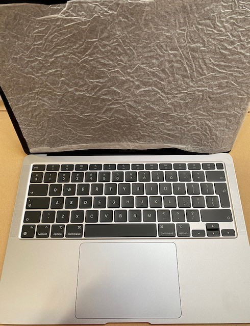 Otwarty srebrny MacBook z czarną klawiaturą. Arkusz ochronny zakrywa górę. Tło to kartonowa powierzchnia.