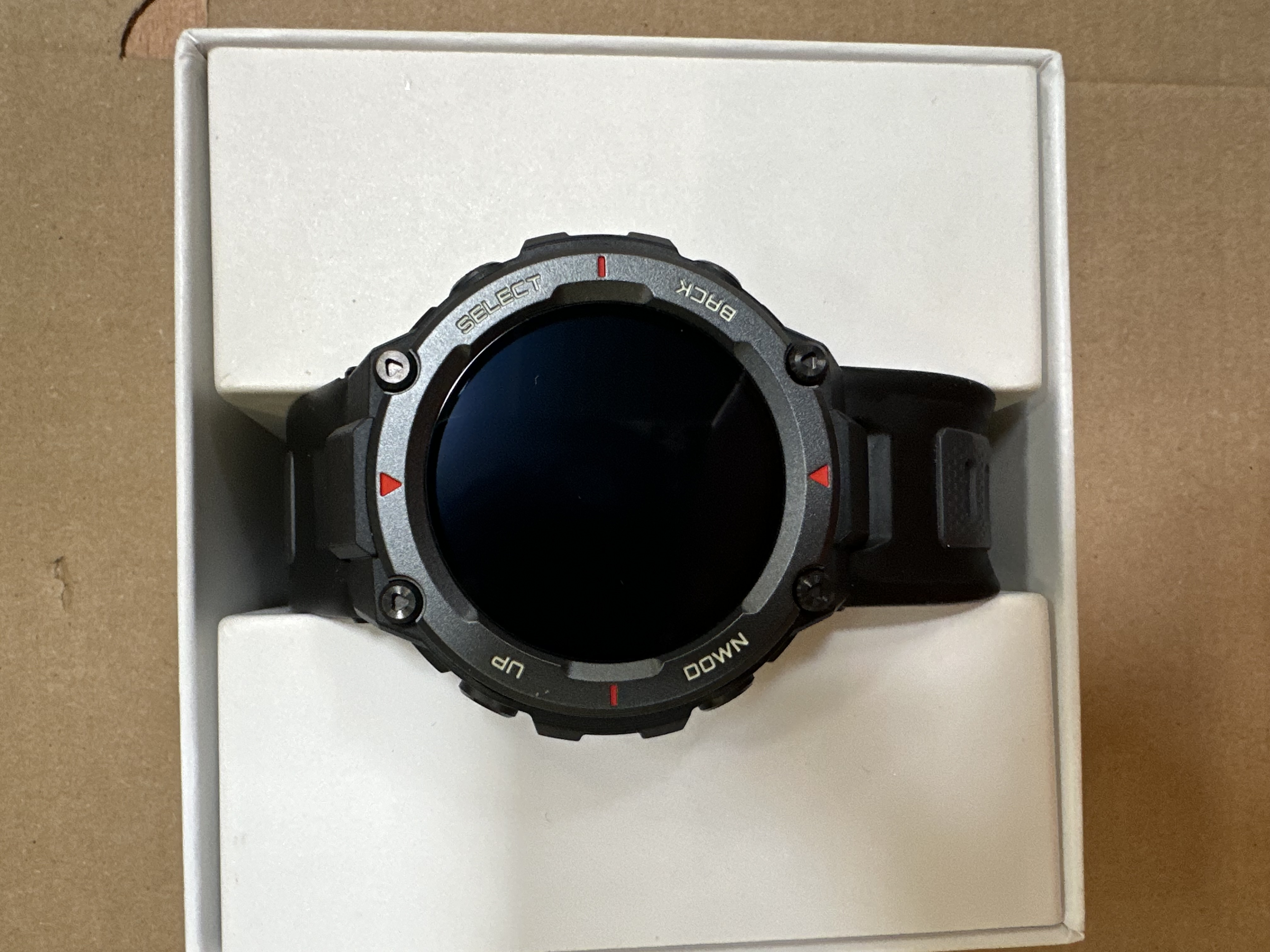 Czarny smartwatch w białym pudełku. Nadrukowane UP, DOWN, SELECT i BACK. Tło brązowej powierzchni.