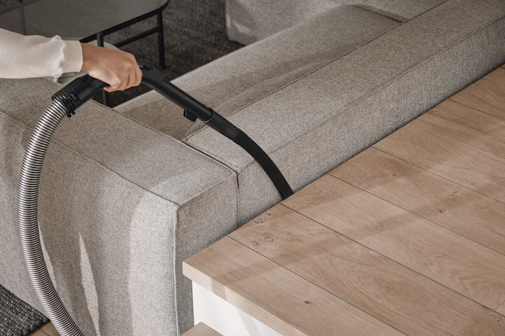 Ręka odkurzająca sofę. Wąż odkurzacza jest widoczny. Drewniany stół po prawej stronie.