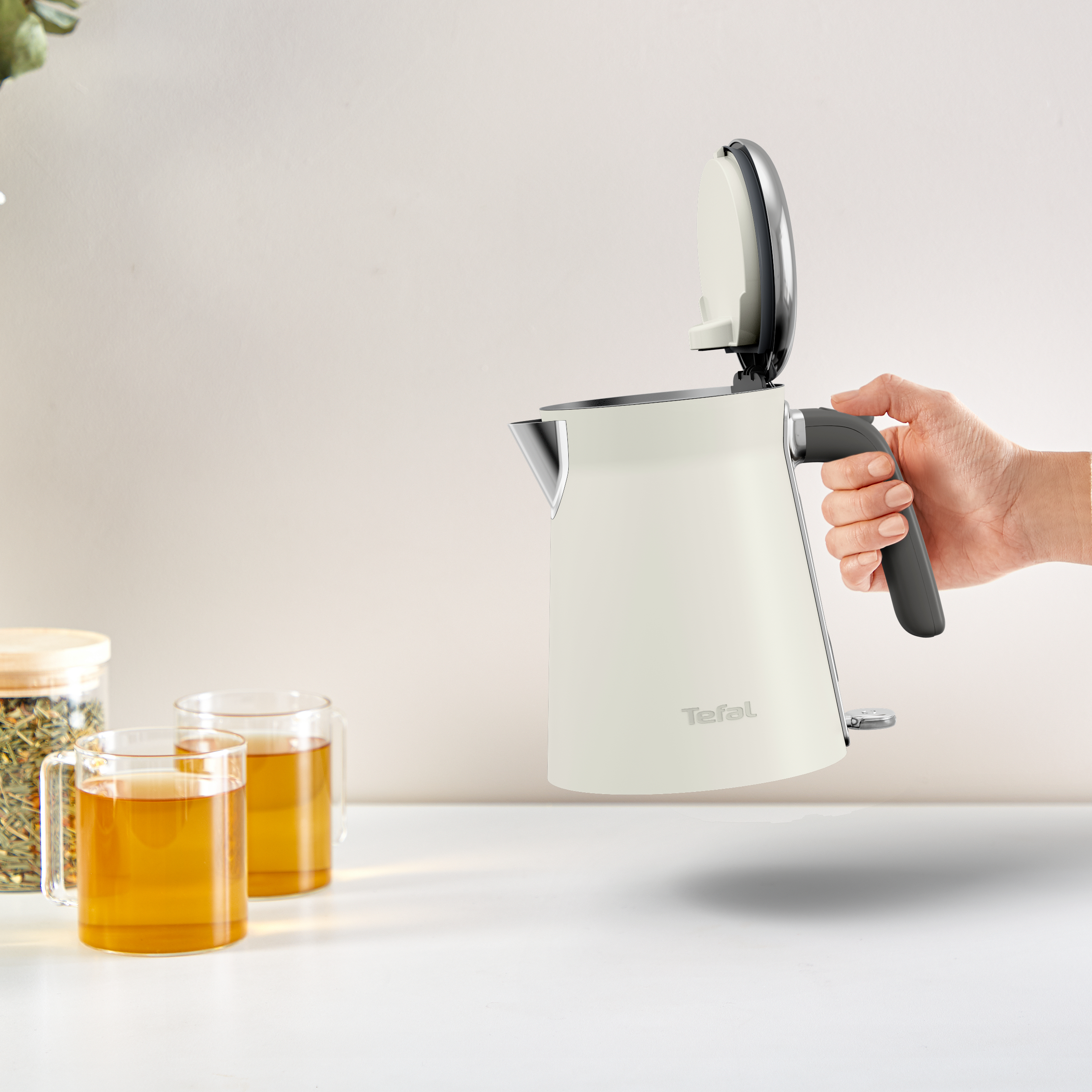 Eine Hand öffnet einen weißen elektrischen Wasserkocher mit grauem Griff. Zwei Tassen Tee sind sichtbar.