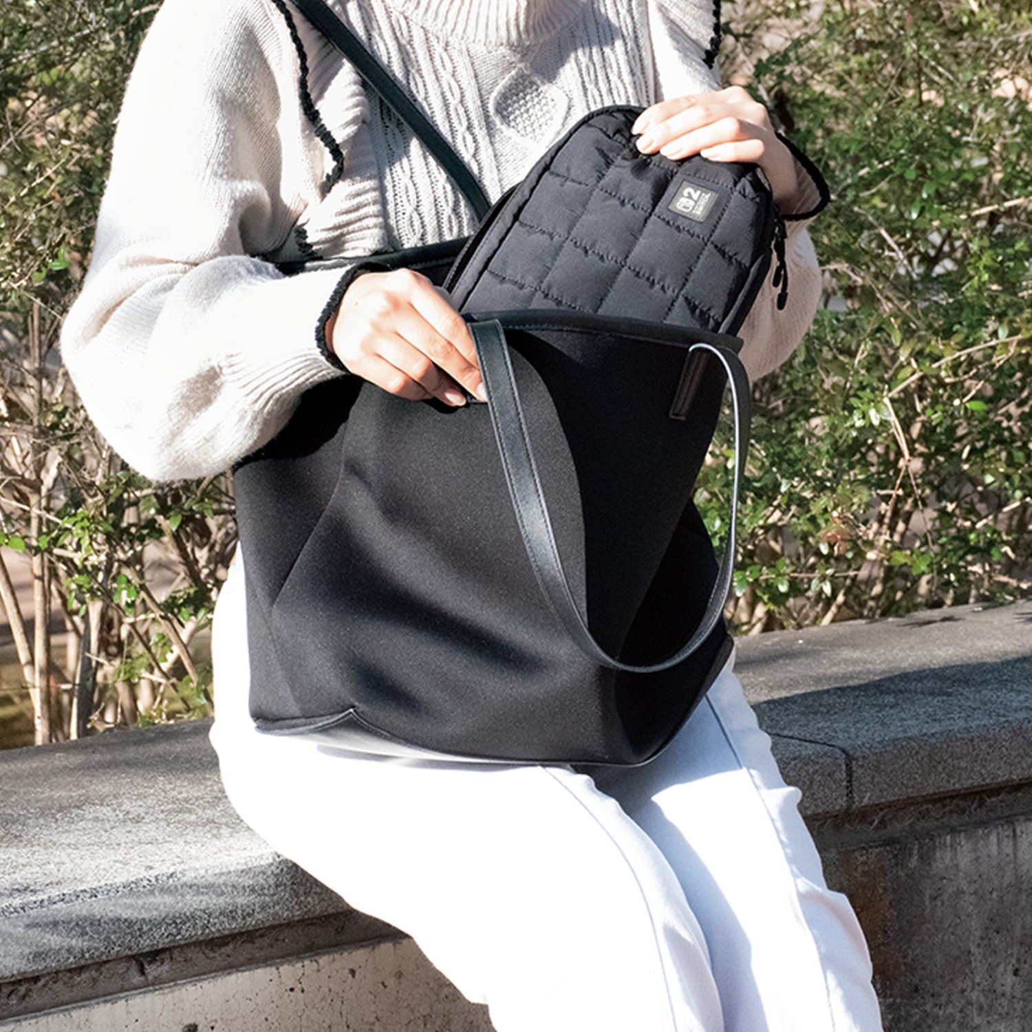 Femme mettant un sac noir matelassé dans un sac fourre-tout noir. En plein air, journée ensoleillée.