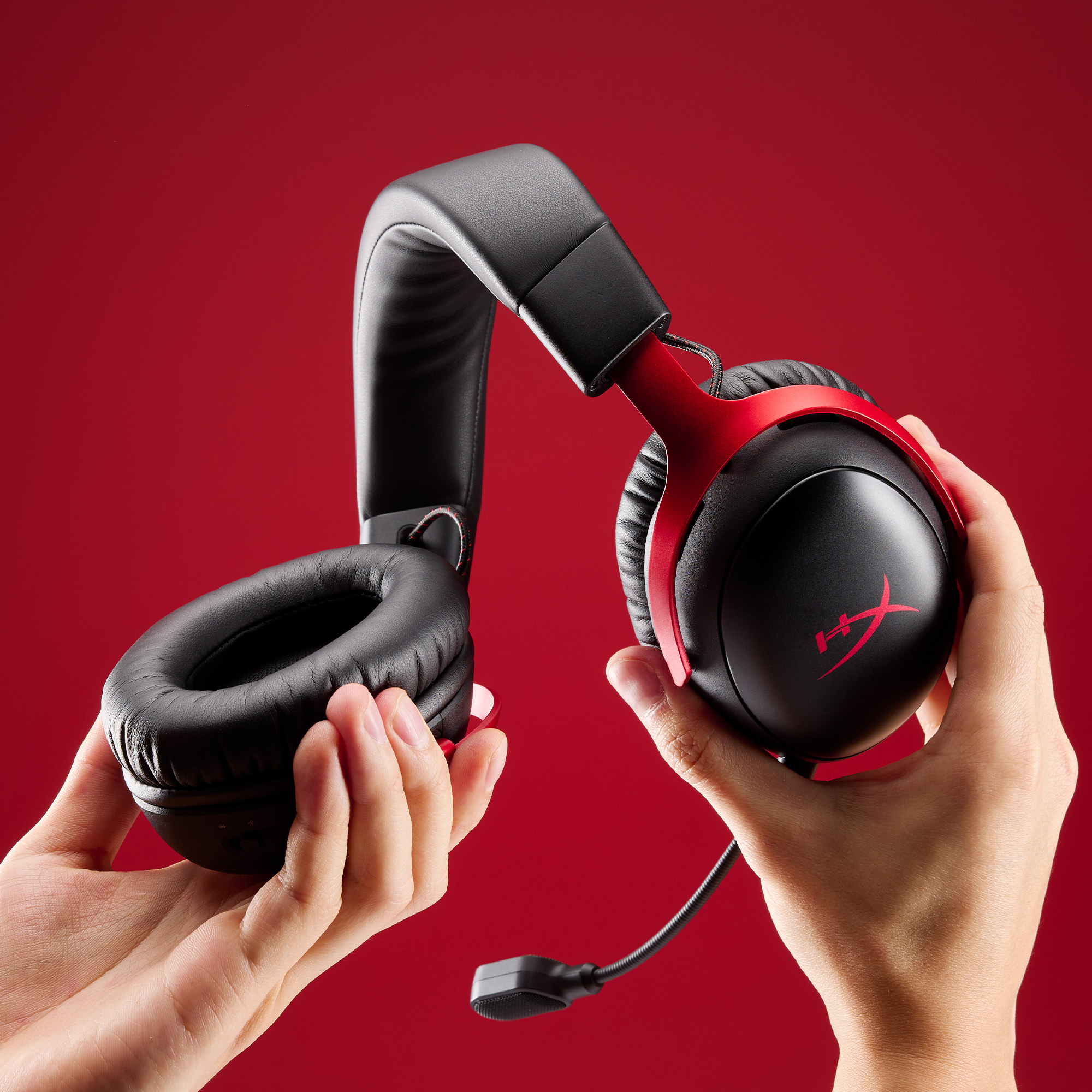 Hände halten schwarzes und rotes HyperX Cloud III Headset mit Mikrofon, roter Hintergrund.