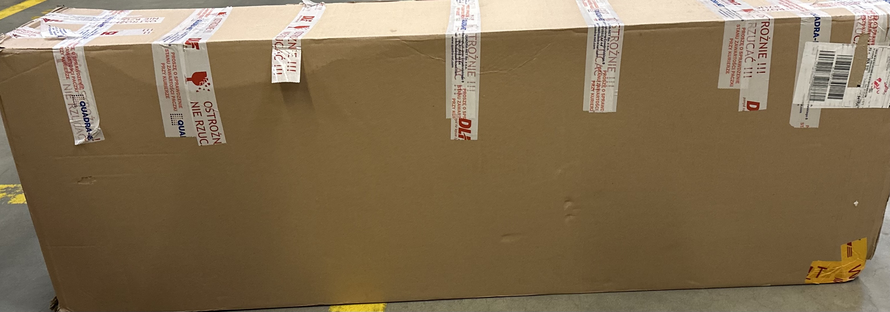 Duże kartonowe pudełko z taśmą i etykietami ostrzegawczymi. Pokazuje DHL i dane wysyłkowe.