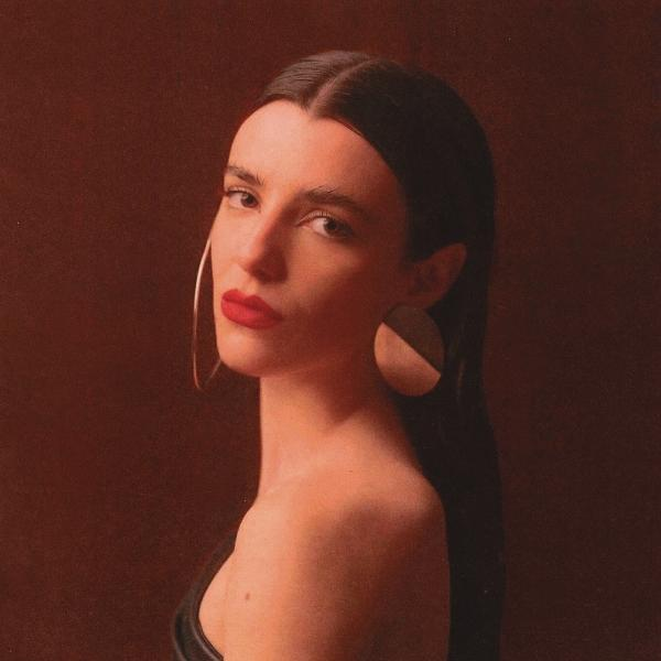 Frau mit dunklem Haar, rotem Lippenstift, großen Ohrringen, brauner Hintergrund.