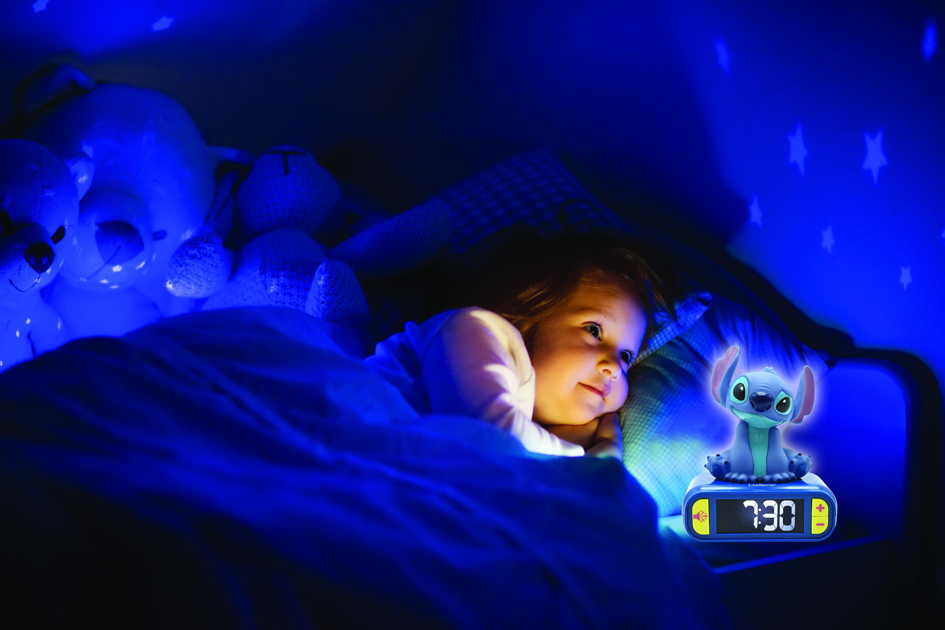Ein Kind liegt nachts im Bett mit einem Stitch-Wecker. Sterne werden an die Wand projiziert. Das Bett ist blau.