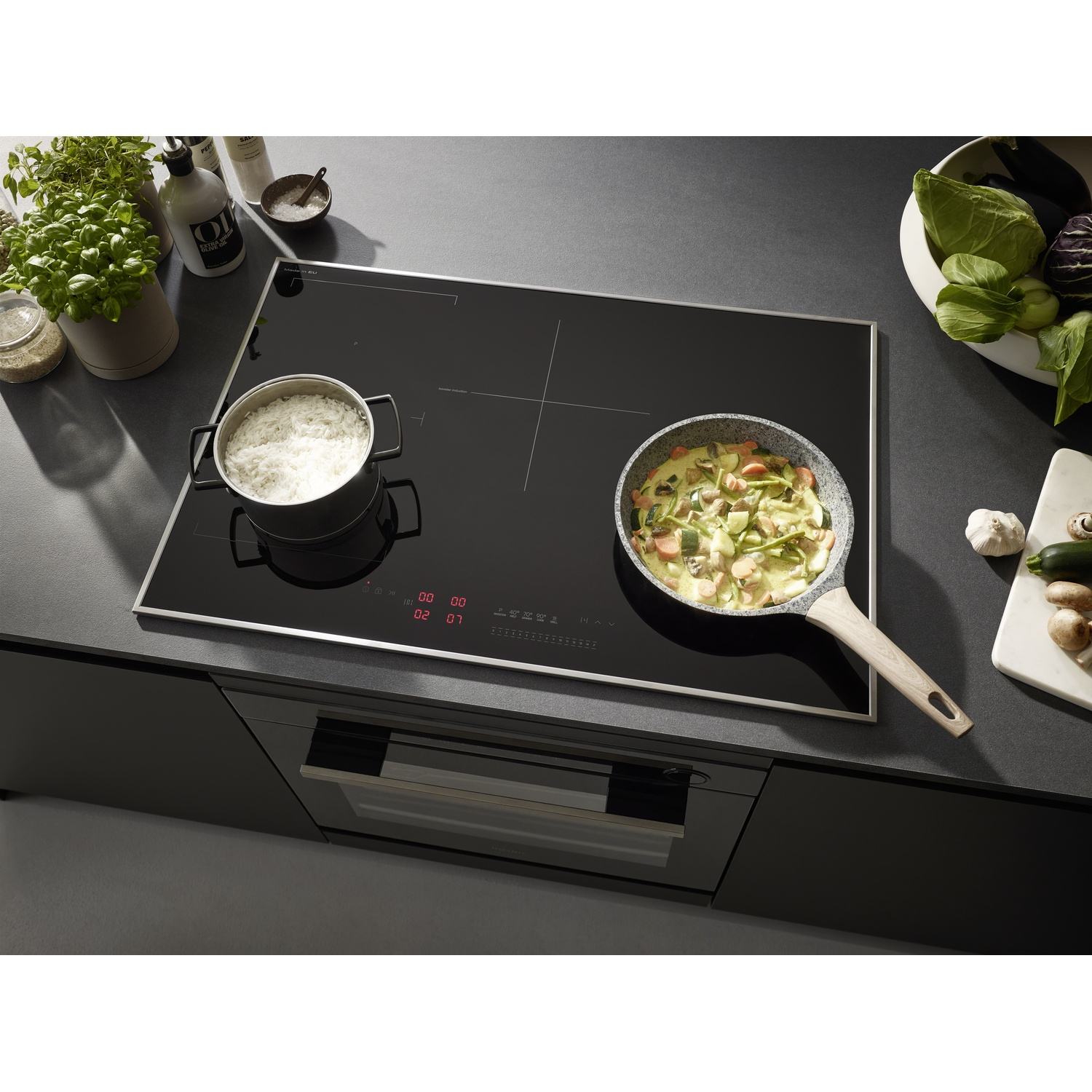 Piano cottura a induzione con pentola e padella con cibo. Piante e utensili da cucina sullo sfondo.