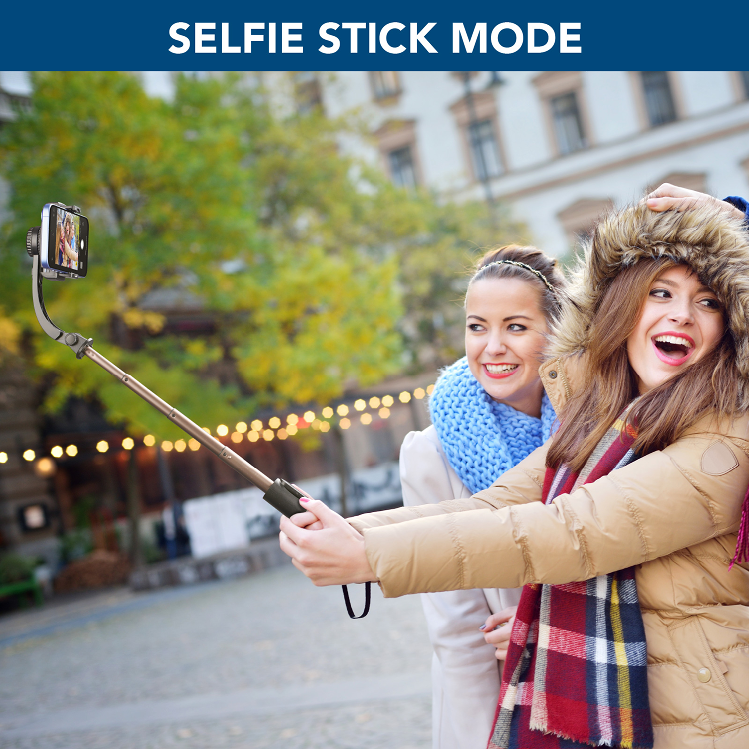 Dos mujeres usando un palo selfie al aire libre. Una sostiene el palo, sonriendo, con abrigo.