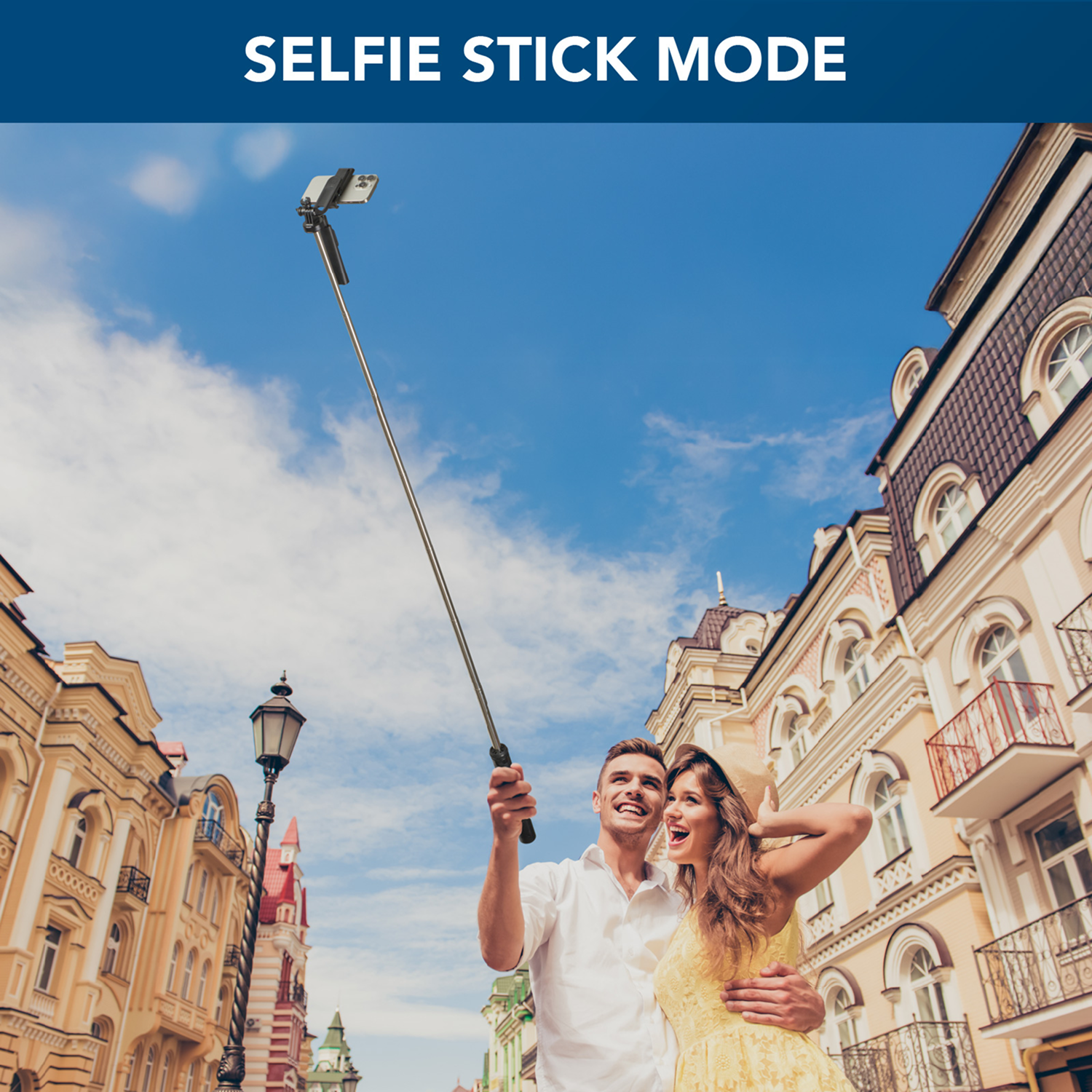 Una pareja usando un palo selfie al aire libre, sonriendo, con un paisaje urbano de fondo.