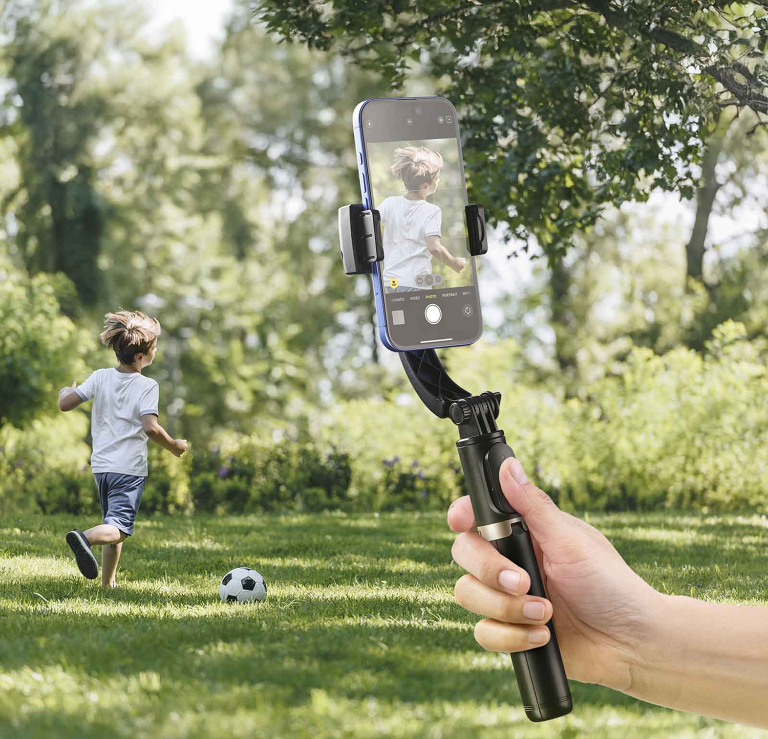 Eine Hand hält einen Selfie-Stick und fängt einen Jungen ein, der mit einem Fußball rennt.