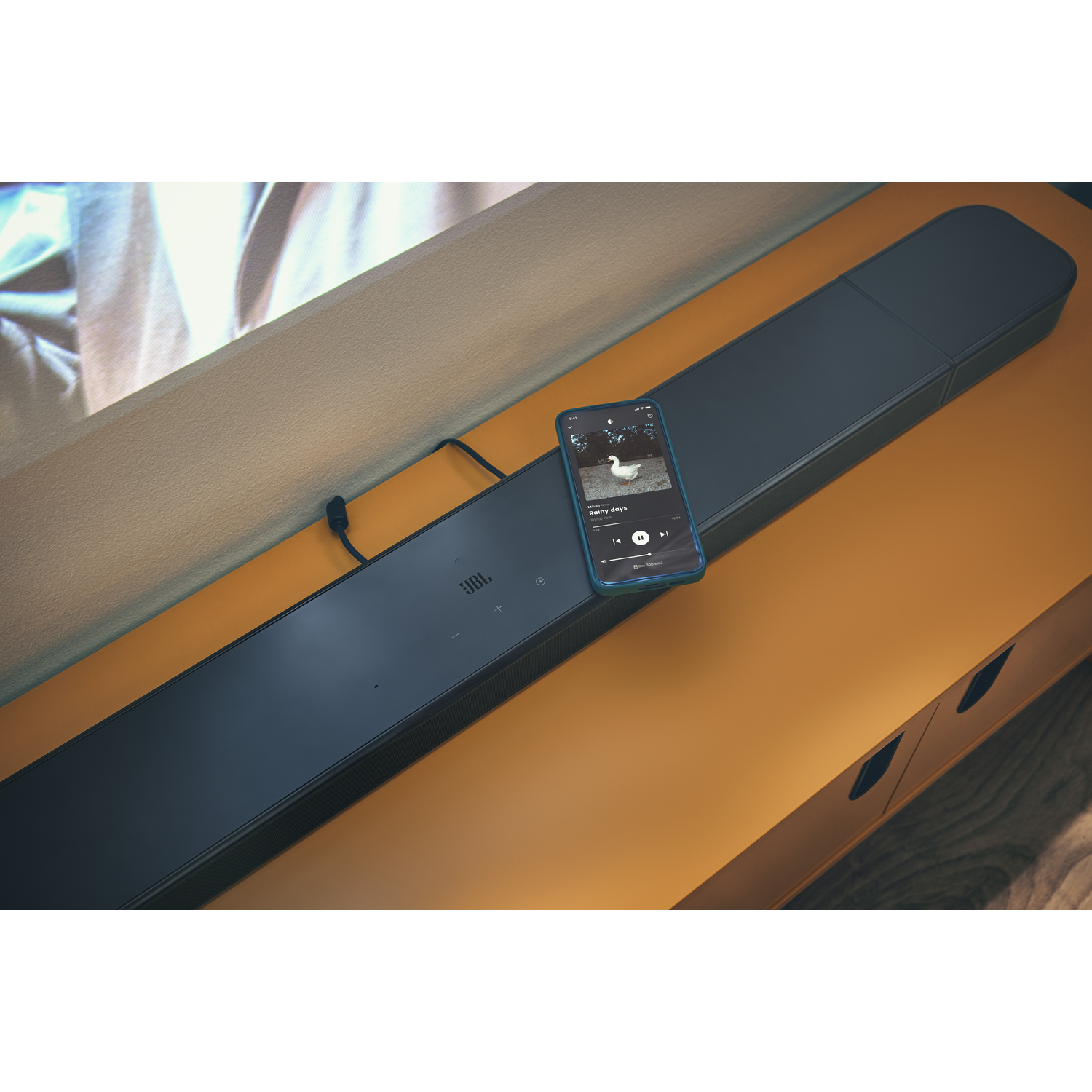 Une barre de son JBL sur un meuble orange. Un téléphone affiche une application musicale.