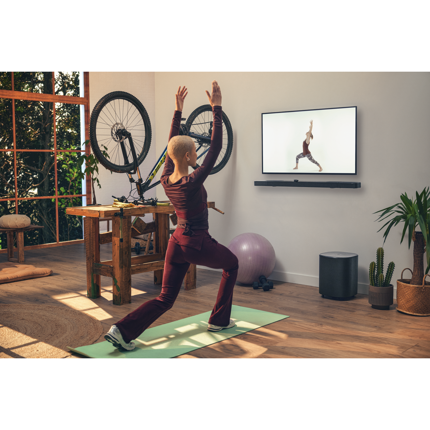 Une femme fait du yoga devant une télévision avec une barre de son. Un vélo est sur le mur.