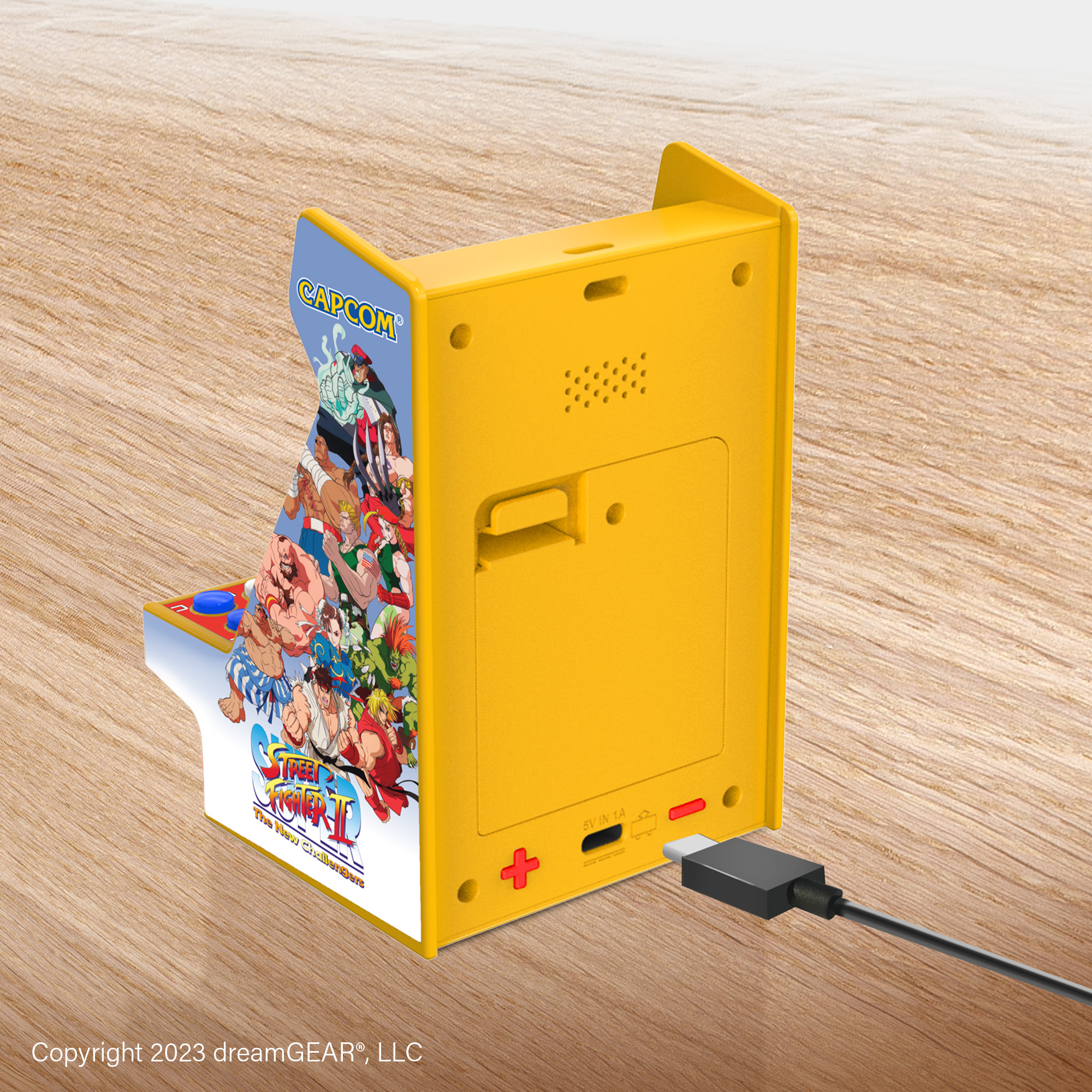 Sarı mini atari oyunu. Bir şarj kablosu yan tarafa takılı. Arkada bir hoparlör var.