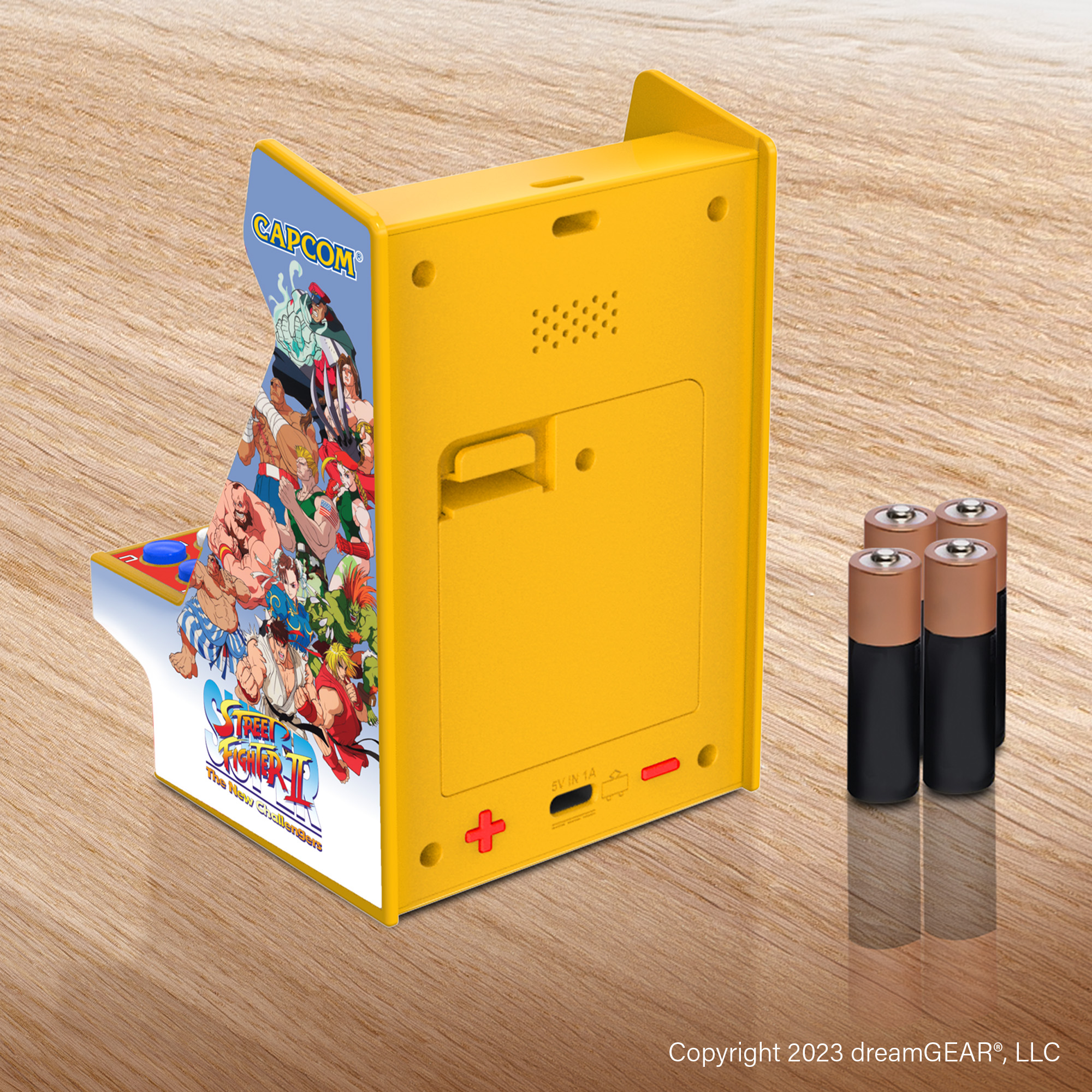Sarı mini atari oyunu. Yanında 3 pil var. Arkada bir hoparlör ve bir şarj portu var.
