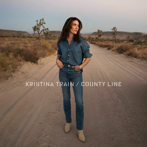 Frau in Jeans steht auf einer unbefestigten Straße in einer Wüstenlandschaft. Der Text lautet: Kristina Train / County Line.