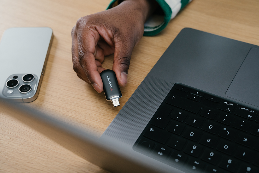 Egy kéz egy Verbatim USB-meghajtót tart egy laptop közelében. Egy telefon fa felületen nyugszik.