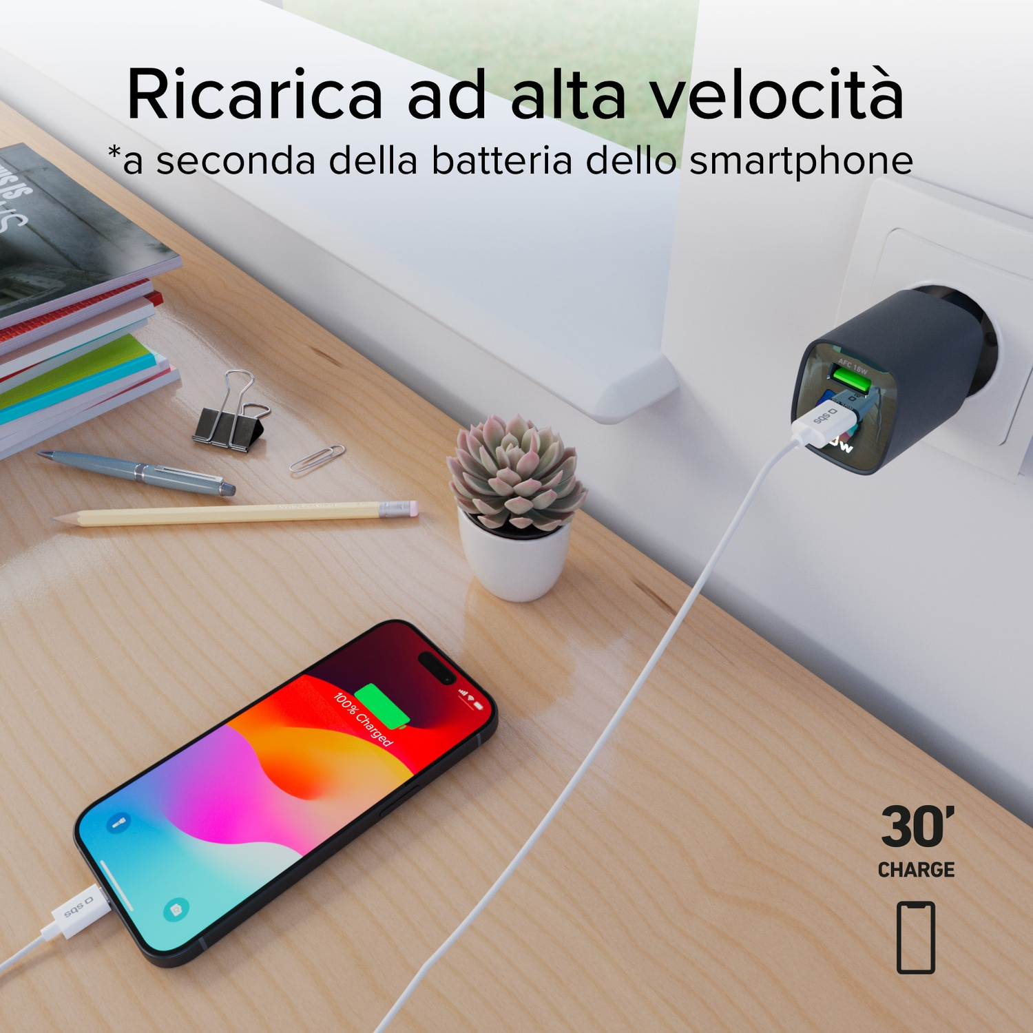 Un telefono che si carica su un tavolo di legno, un caricatore in una presa a muro. Sono visibili una pianta e cancelleria.