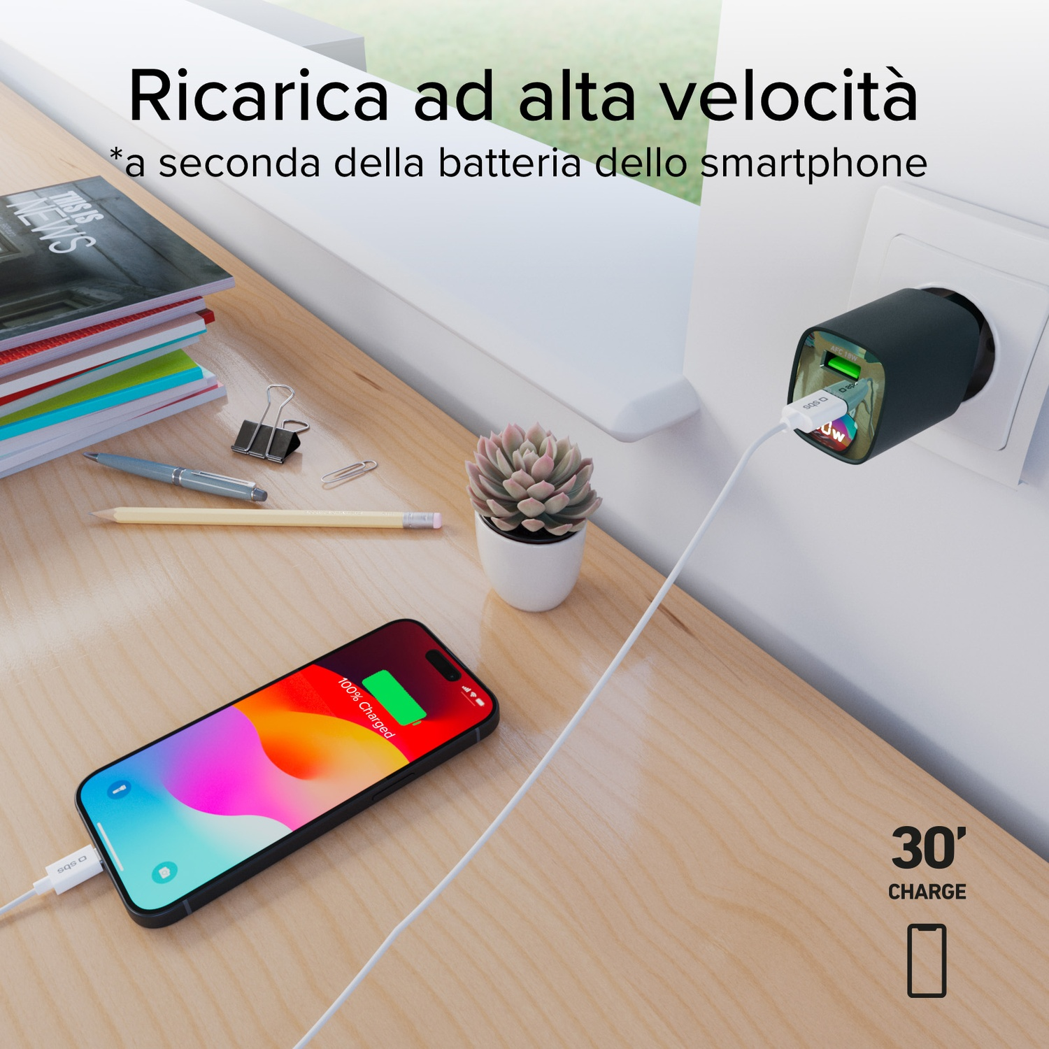 Un telefono che si carica su un tavolo di legno, un caricatore in una presa a muro. Sono visibili una pianta e cancelleria.
