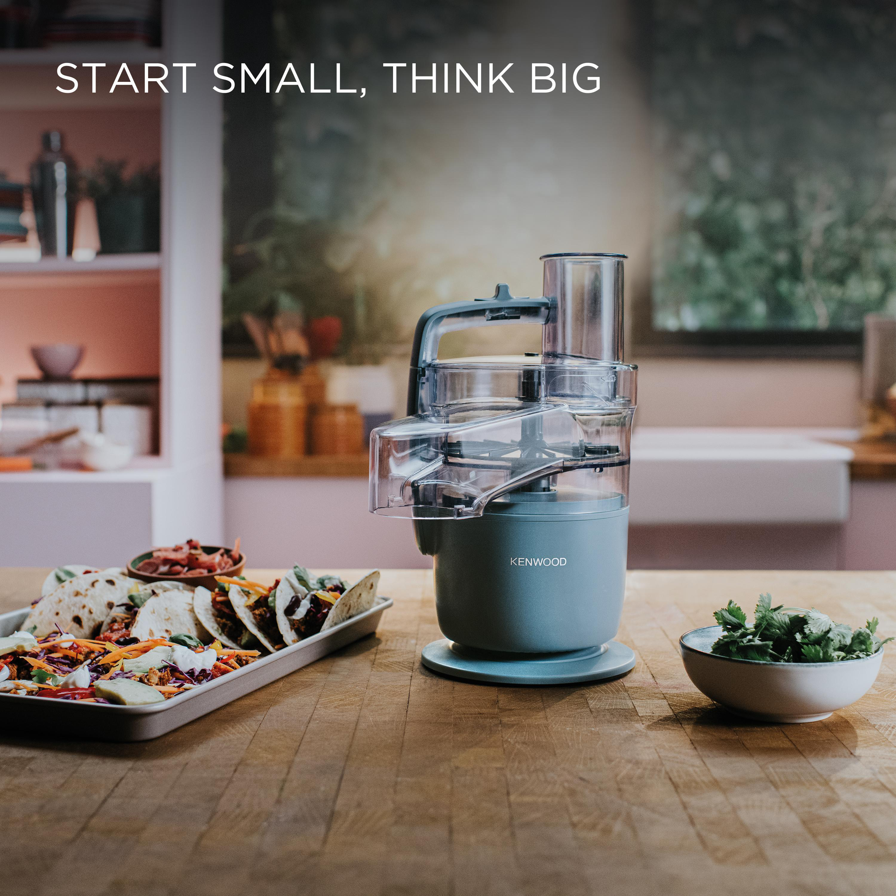 Küchenmaschine auf Holztisch, Tacos, Kräuterschüssel. Text: START SMALL, THINK BIG. Hintergrund ist unscharf.