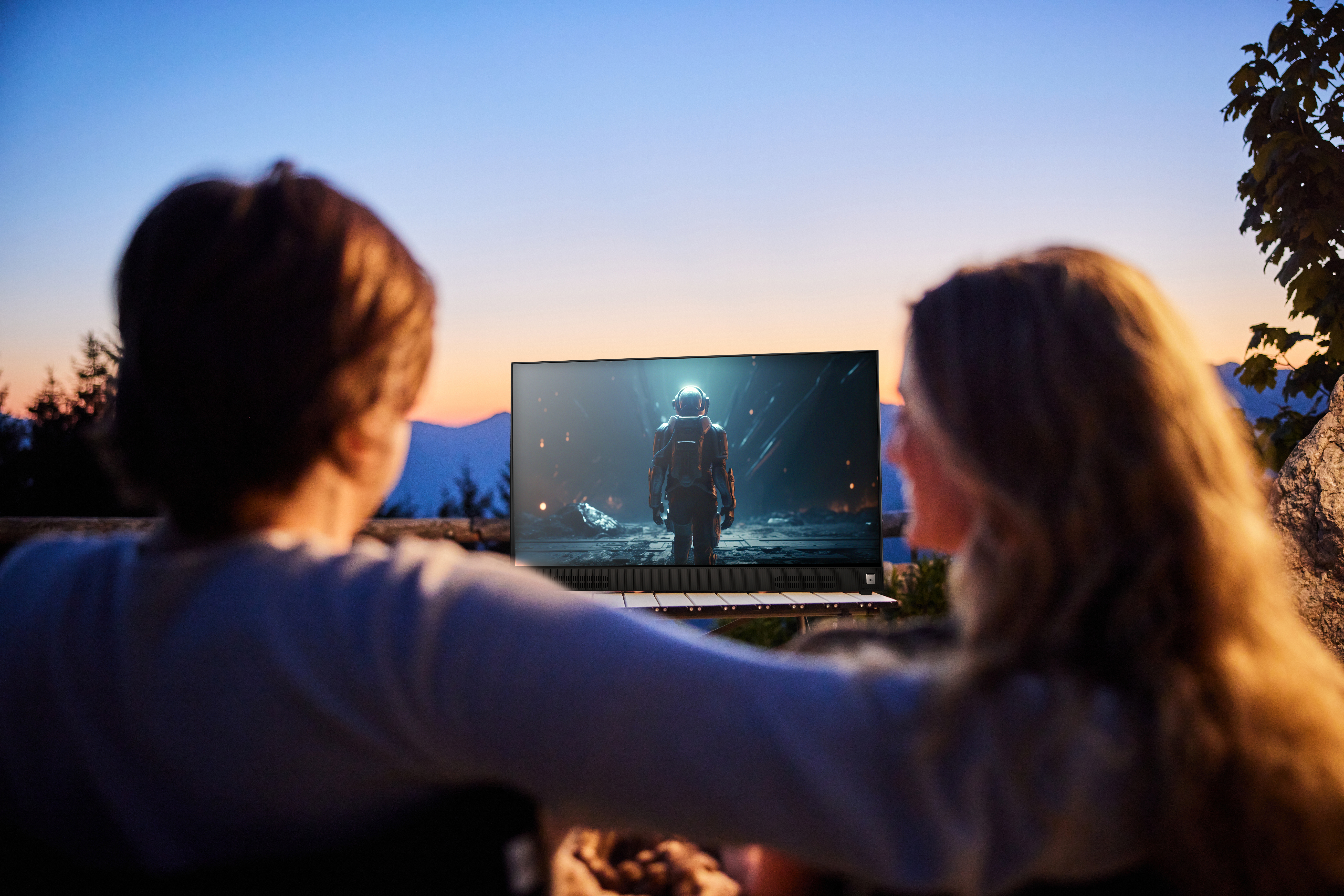 Paar, das im Freien bei Sonnenuntergang eine Sendung im Fernsehen anschaut, Berge im Hintergrund.