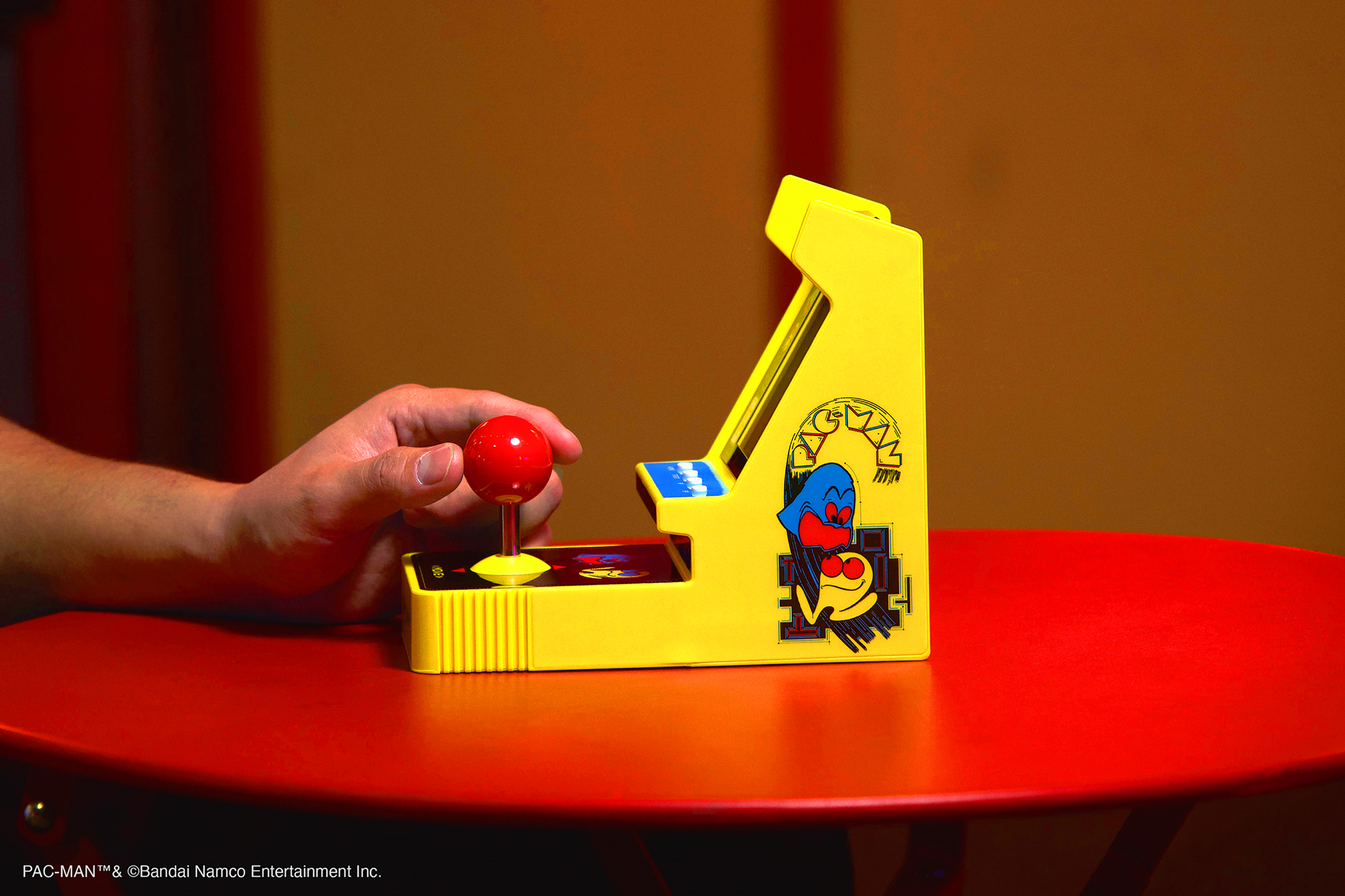 Kırmızı masada sarı atari makinesi. Kırmızı joystick ve Pac-Man grafikleri görünür. Solda bir adamın eli.