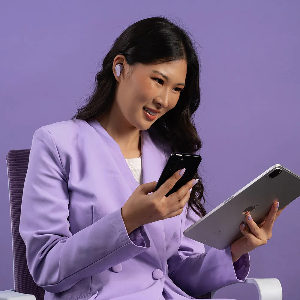 Donna in giacca viola con telefono e tablet, con auricolari, sorridente. Sfondo viola.