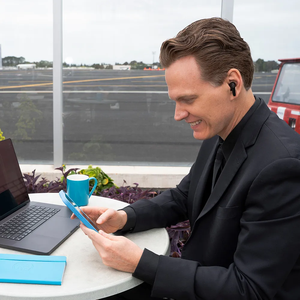 Mann im Anzug mit Ohrhörern, telefoniert, lächelt. Laptop, blauer Becher und Notizbuch auf dem Tisch.