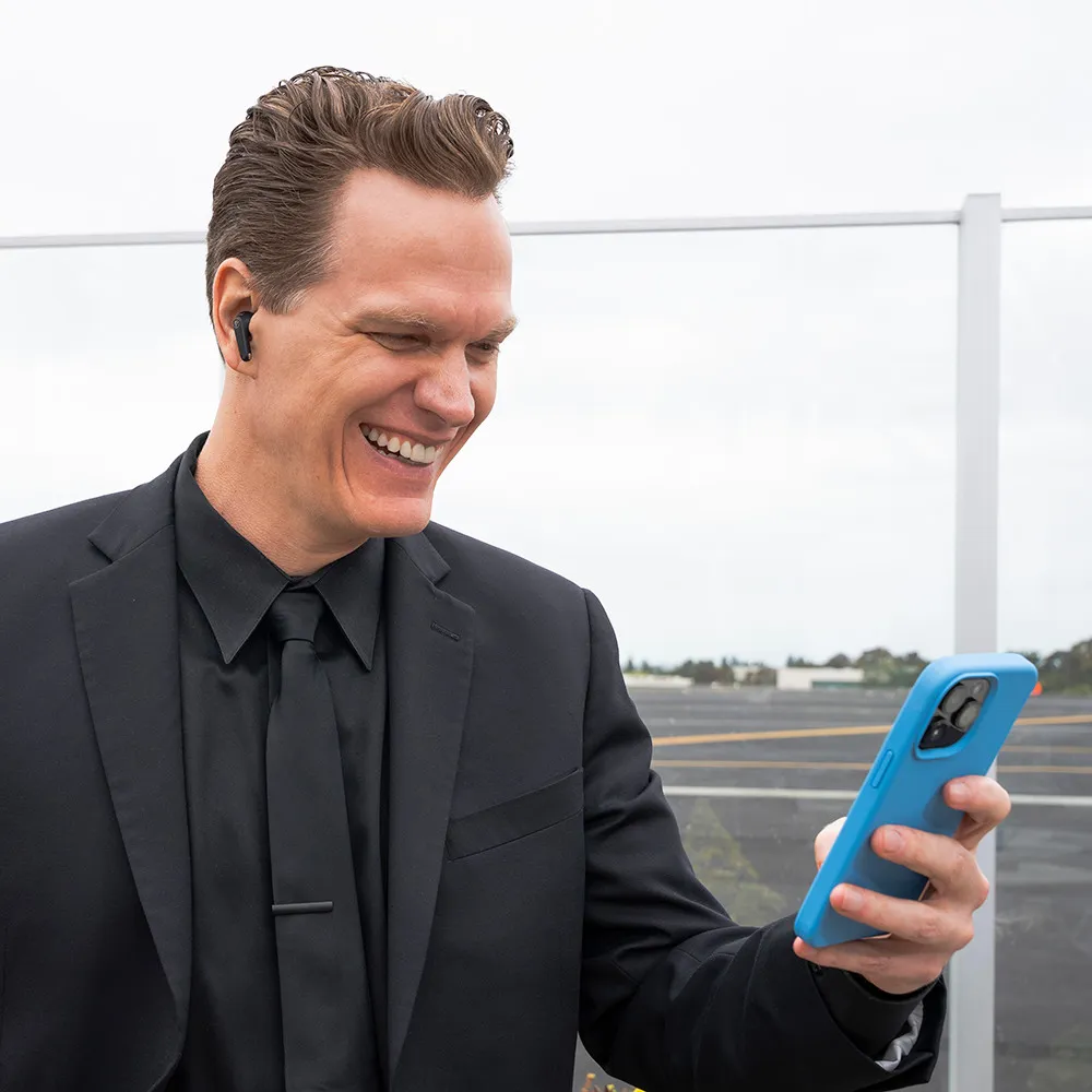 Mann im Anzug mit Ohrhörern, schaut auf ein Telefon, lächelt. Ein Flughafen ist im Hintergrund.