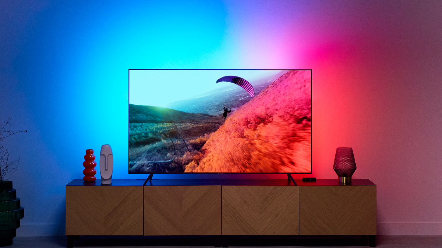 Televisor con escena de parapente, con iluminación de pared azul y roja, sobre un mueble de madera.