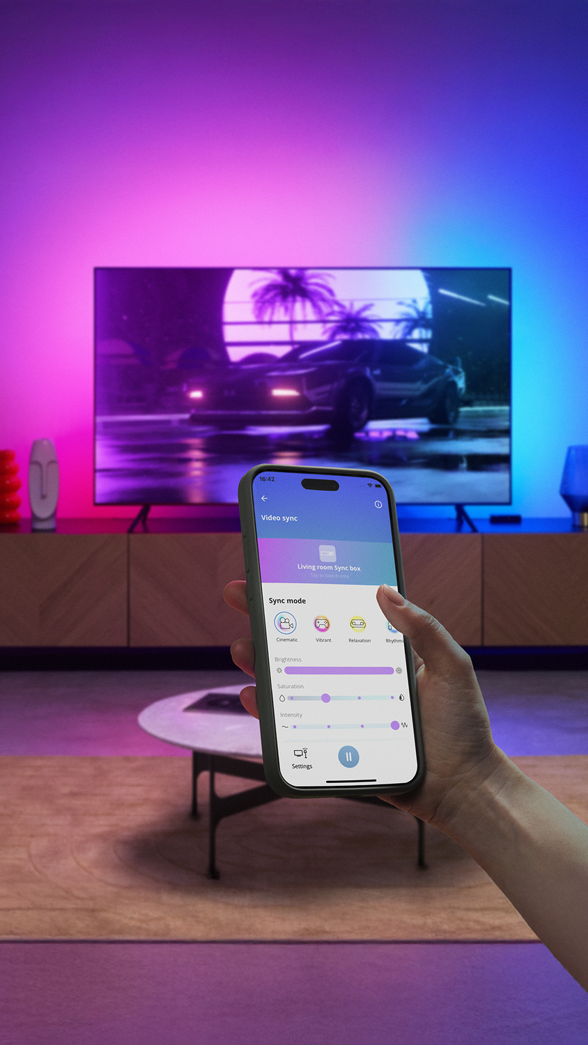 Mano sosteniendo un teléfono que muestra la app WiZ controlando luces detrás de un televisor con escena de coche.