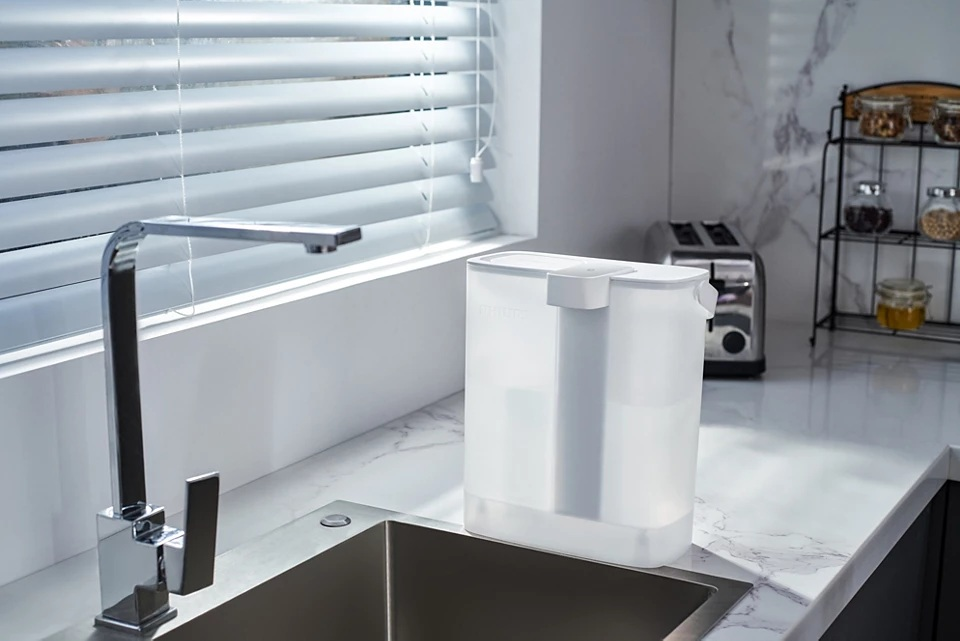 Scena kuchenna: filtr do wody, kran i zlew na pierwszym planie. Toster i przyprawy w tle, w pobliżu okna.