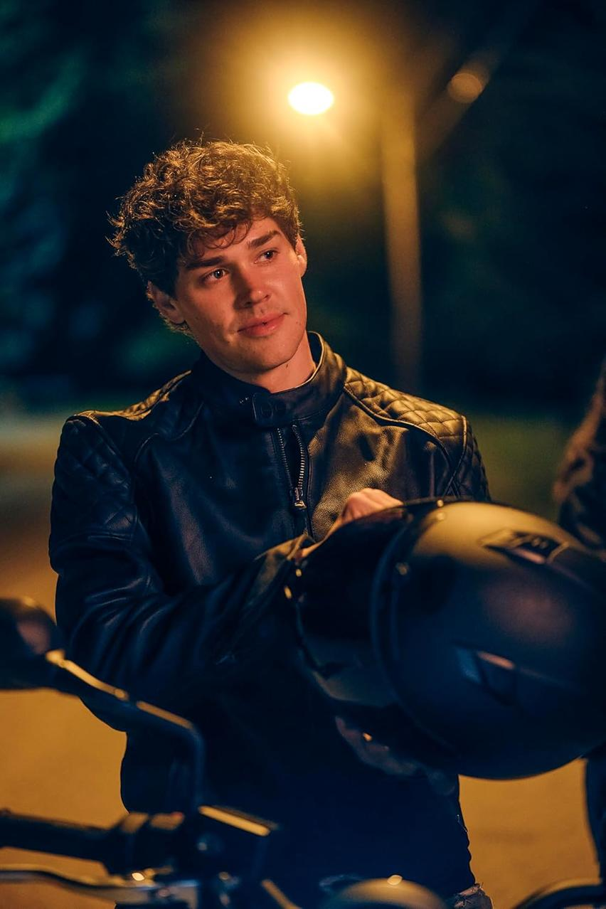 Ein Mann in einer schwarzen Lederjacke hält einen Helm in der Nähe eines Motorrads bei Nacht unter einer Straßenlaterne.
