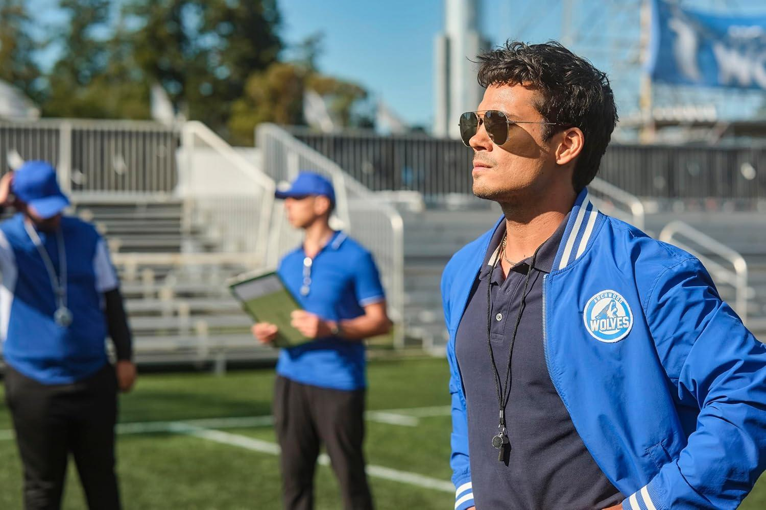 Ein Trainer mit Sonnenbrille steht auf einem Fußballfeld und beobachtet. Zwei weitere Trainer sind im Hintergrund.