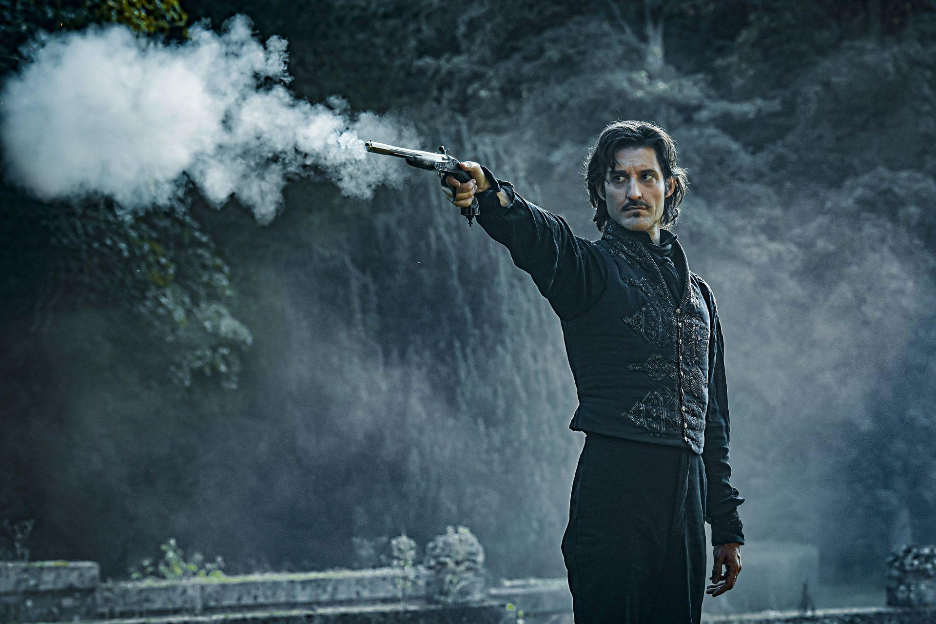 Mann in schwarzem Kostüm feuert eine Pistole ab, Rauch quillt hervor. Bäume im Hintergrund.