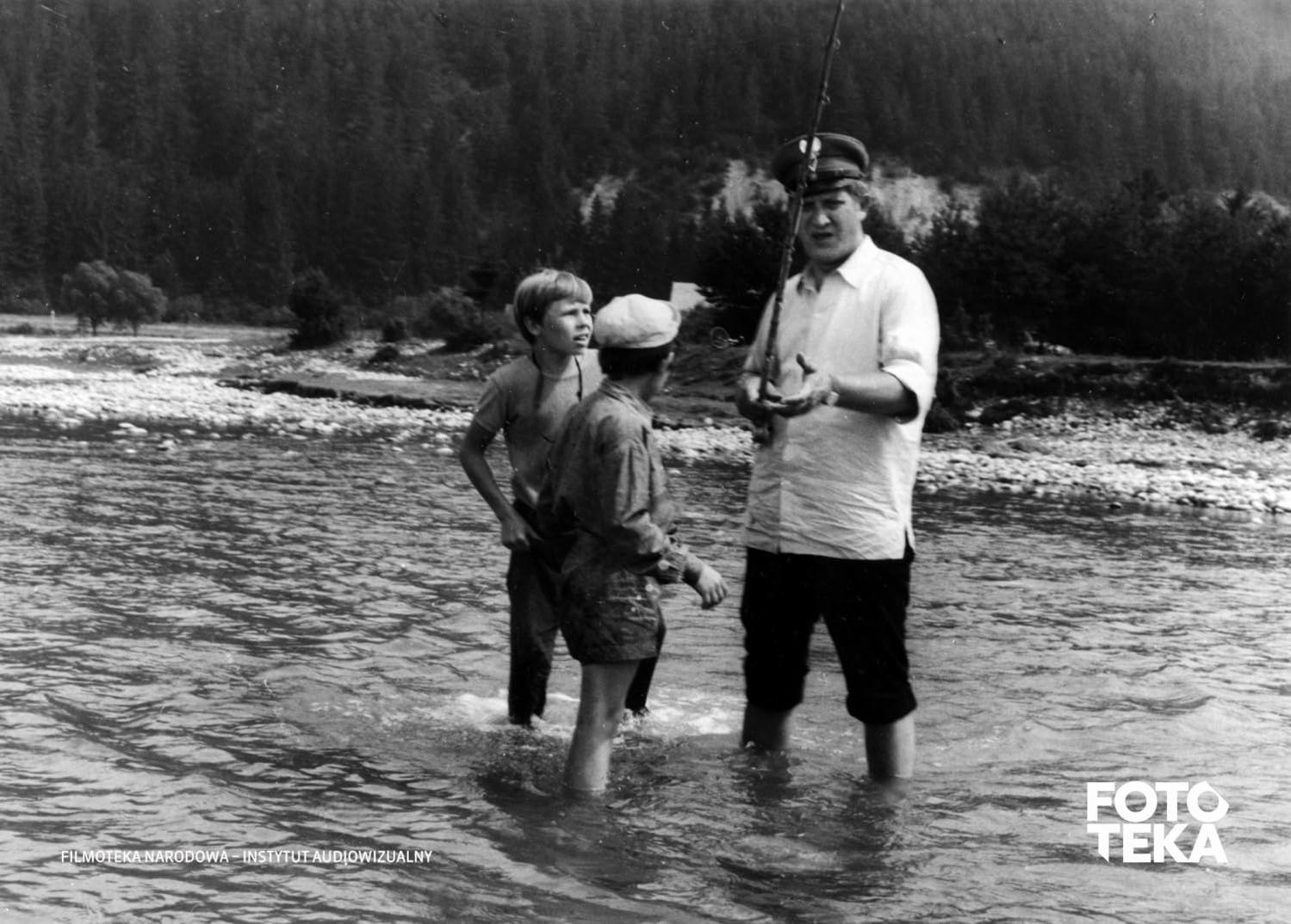 Drei Jungen und ein Mann angeln in einem Fluss. Sie stehen im Wasser, Bäume im Hintergrund.