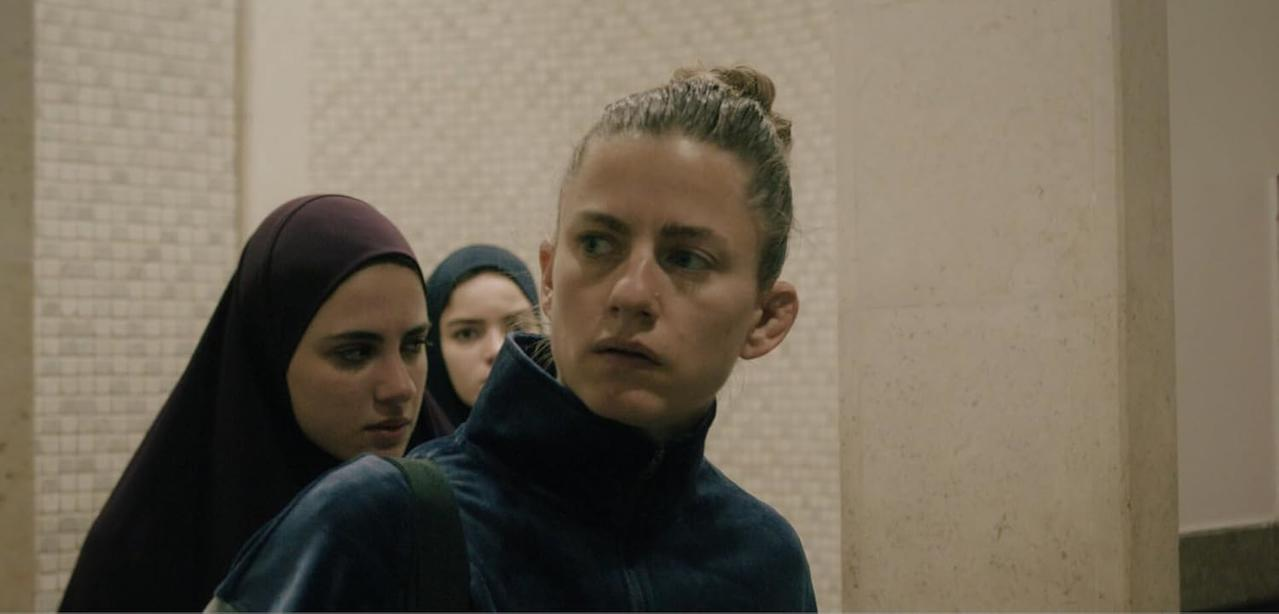 Drei Frauen mit Kopftüchern gehen einen Flur entlang. Eine schaut nach vorne, andere zur Seite.