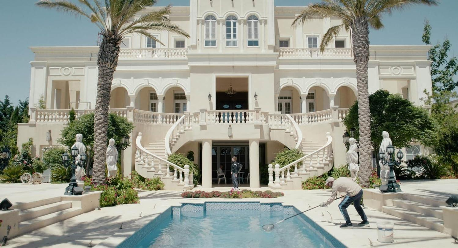 Eine große weiße Villa mit Pool. Zwei Palmen flankieren das Gebäude. Eine Person reinigt den Pool.
