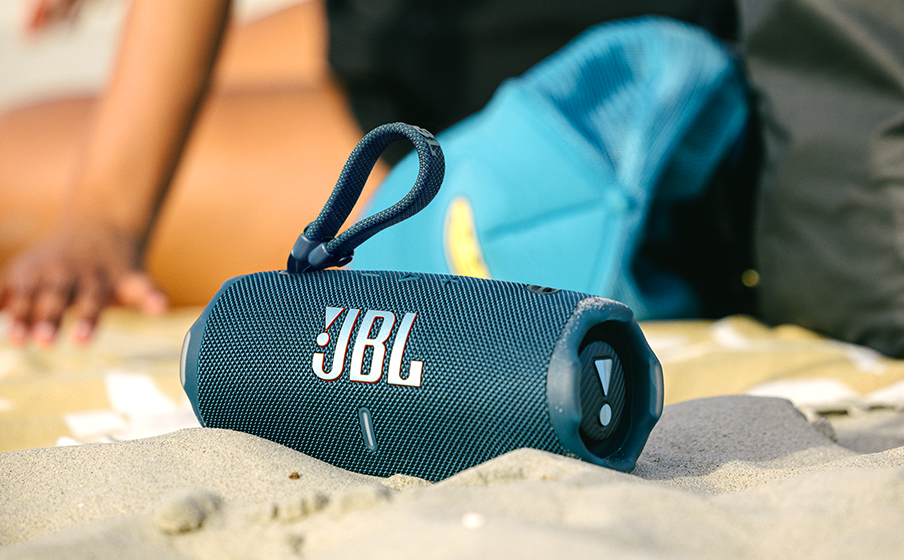 Altavoz JBL azul en arena; persona y sombrero en el fondo. Logotipo visible.