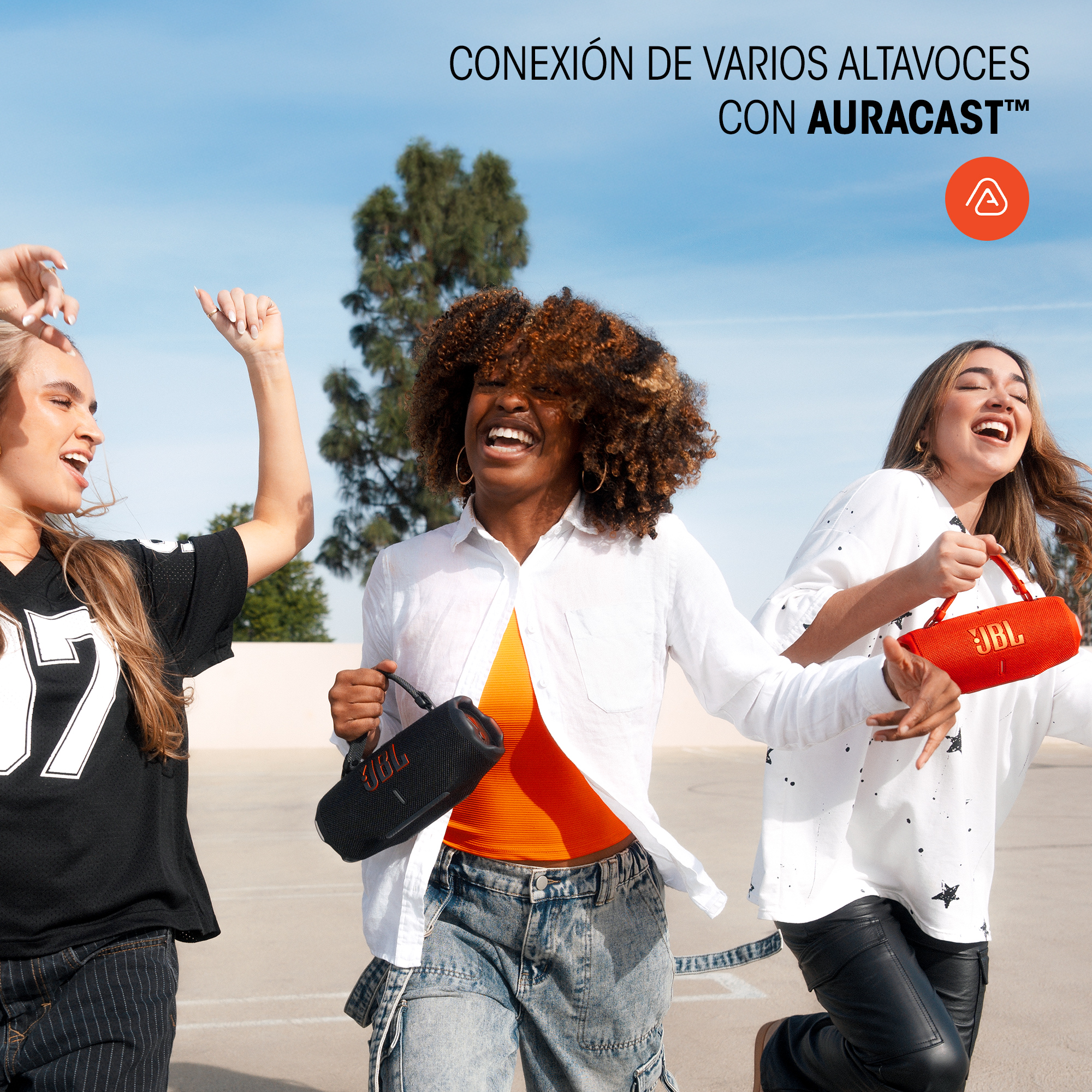 Tres mujeres corriendo, sosteniendo altavoces JBL. Cielo azul, día soleado. Sonrisas.