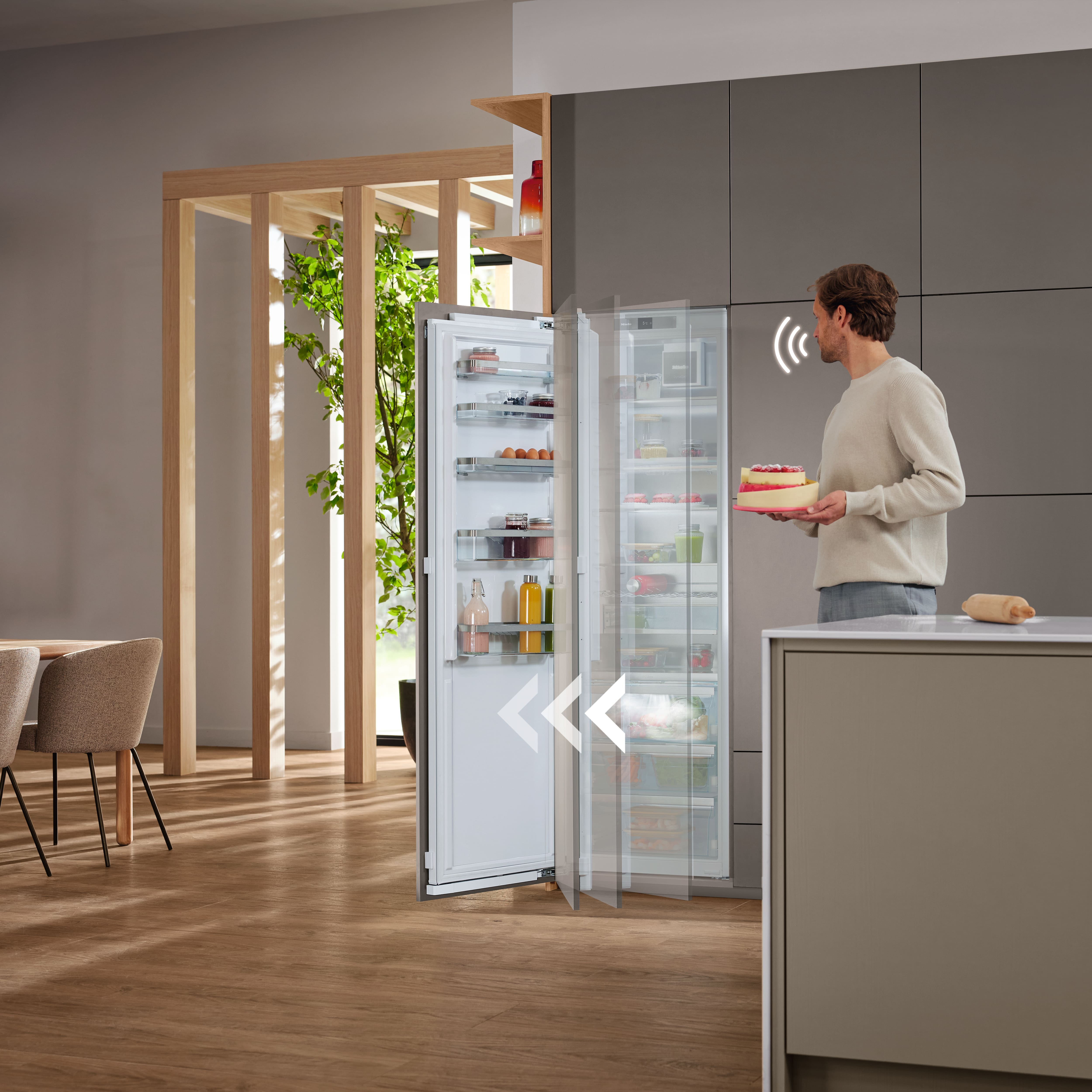 Eine Person mit Kuchen steht vor einem offenen Miele-Kühlschrank. Küche und grüne Pflanzen im Hintergrund.