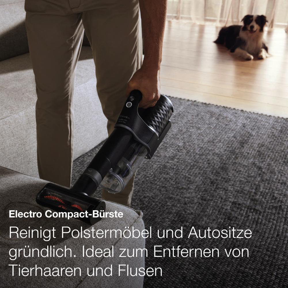 Staubsauger reinigt Sofa. Person hält ihn. Hund im Hintergrund.