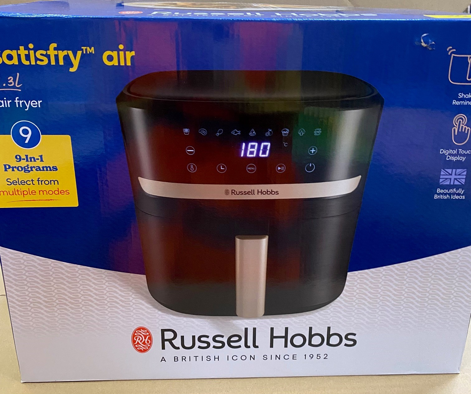 Czarna frytkownica beztłuszczowa Russell Hobbs z cyfrowym wyświetlaczem. Stoi na niebiesko-białym pudełku. Pudełko mówi 'Satisfry air'.