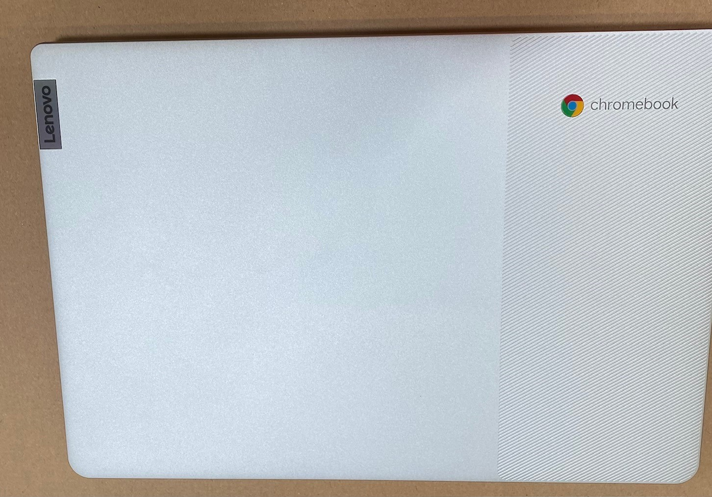 Biały laptop Lenovo Chromebook. Pokrywa ma logo Lenovo i logo Chromebook. Jest na kartonowej powierzchni.