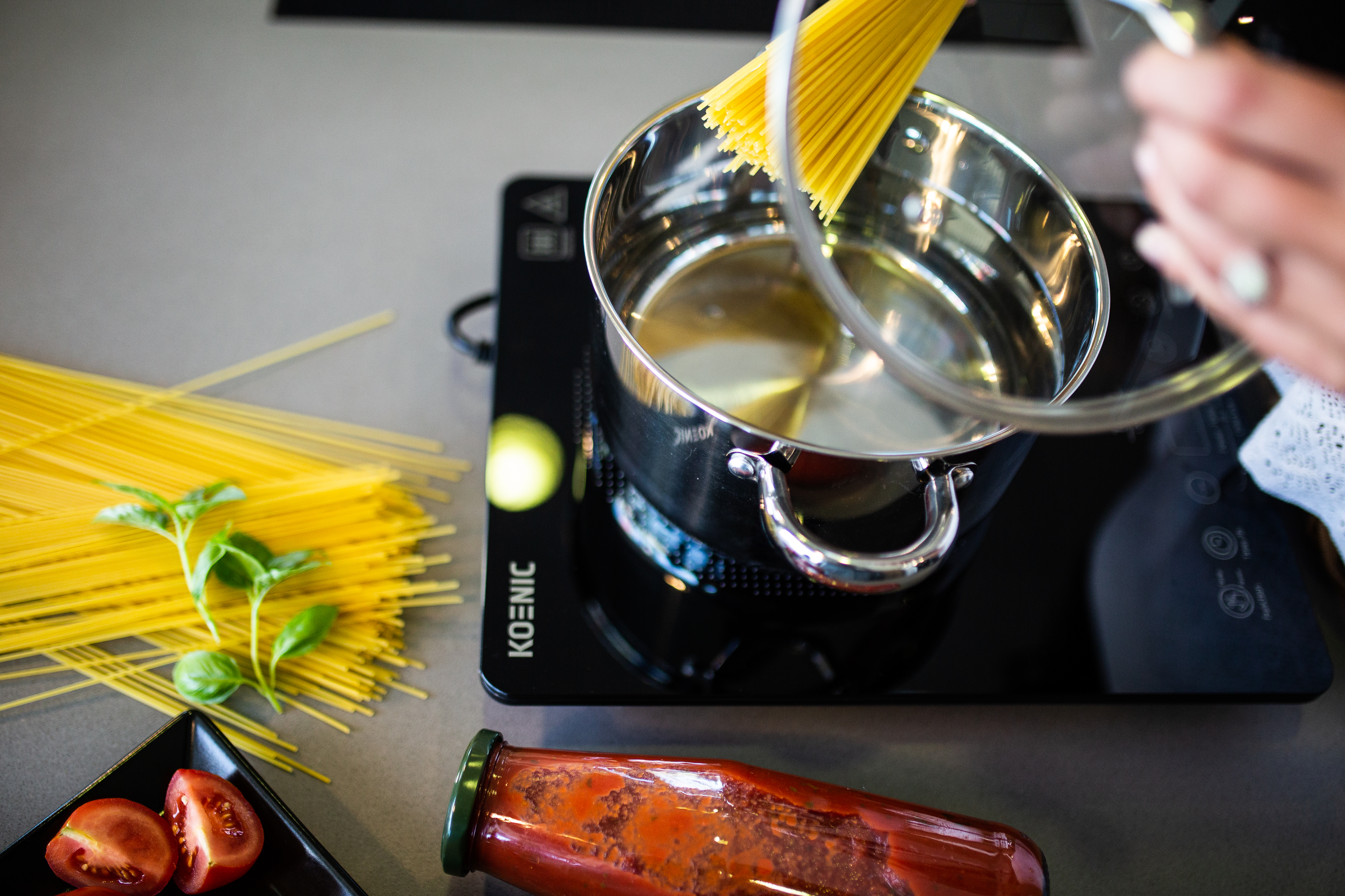 Spaghetti wird in einen Topf mit kochendem Wasser gegeben, mit Sauce, Tomaten und Basilikum daneben.