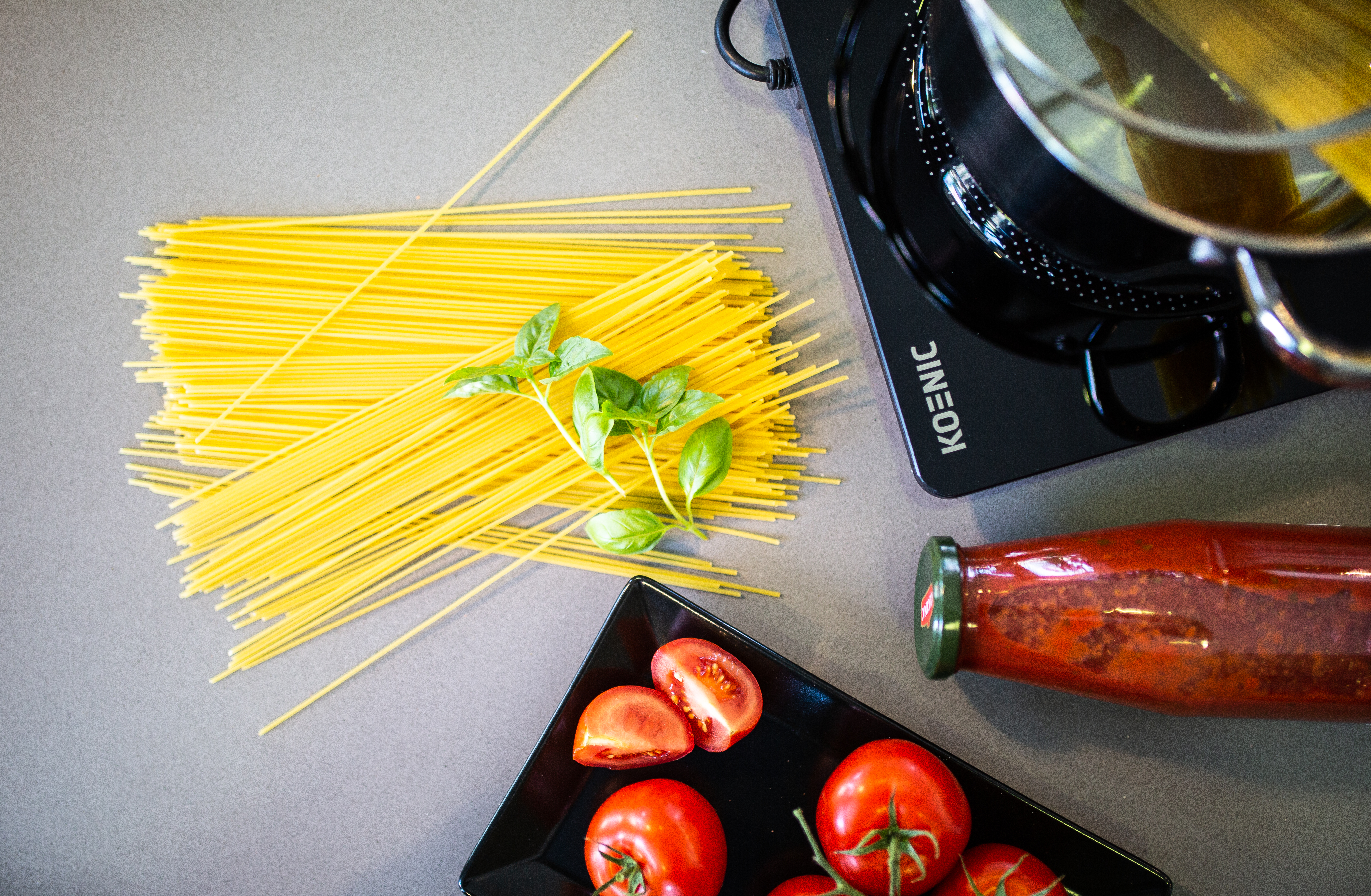 Spaghetti, Basilikumblätter und Tomatenscheiben auf grauer Oberfläche. Schwarzer Topf und Glas daneben.